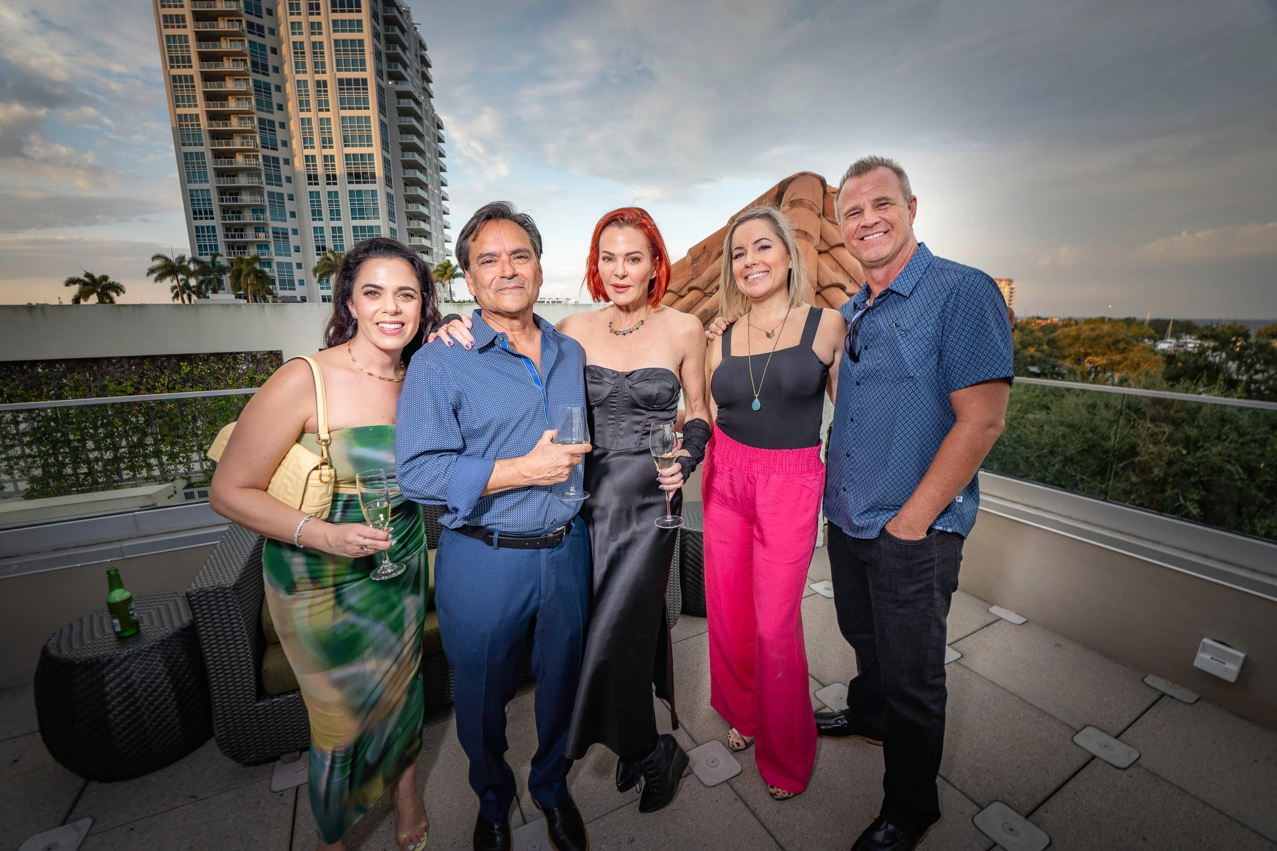 Group of five people at a rooftop party, standing close together with drinks, smiling, with a cityscape and partly cloudy sky in the background.