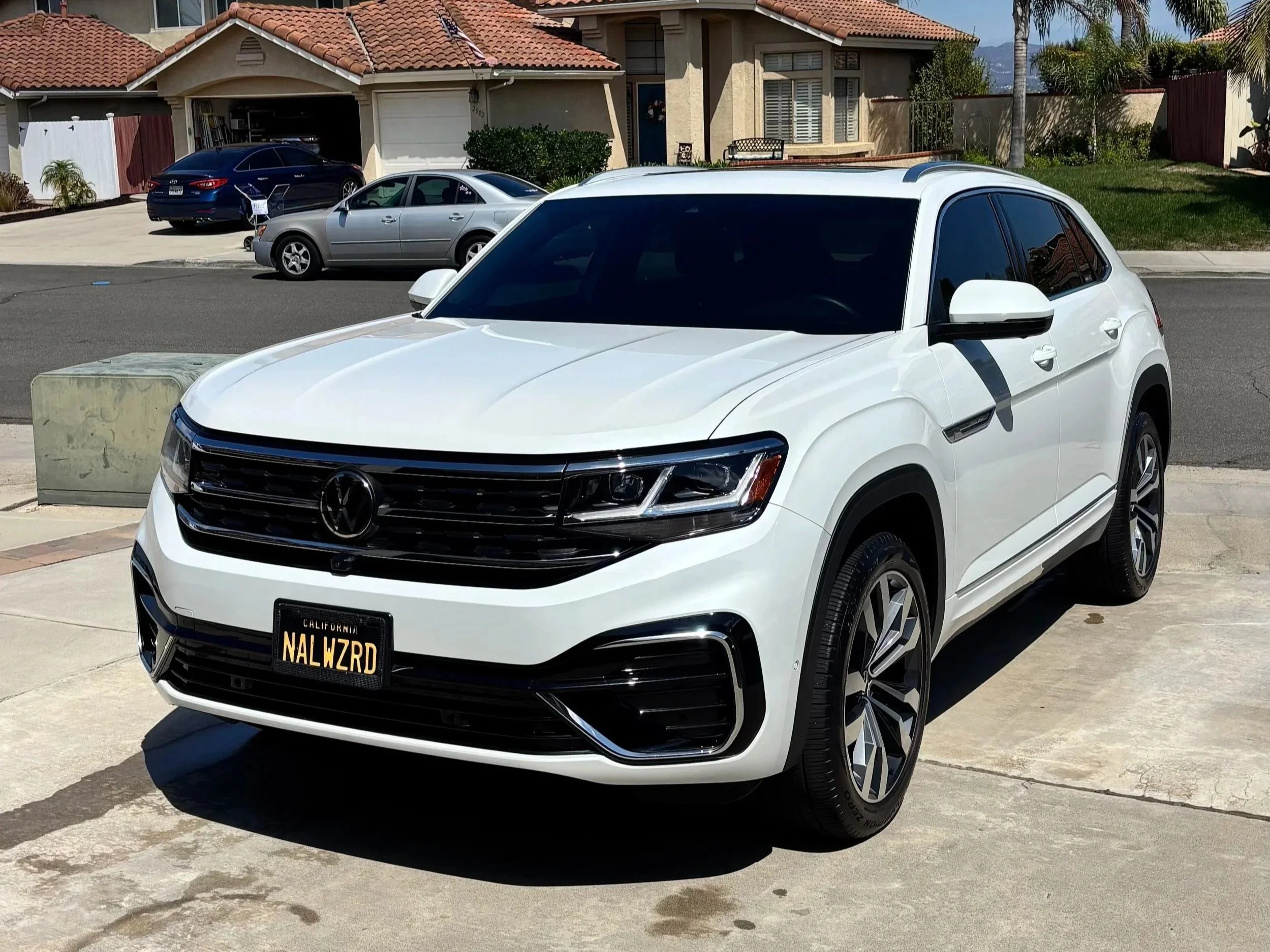 White Volkswagen SUV parked on a driveway in front of a house with a red tile roof, other cars parked across the street, with palm trees and a clear blue sky in the background.
