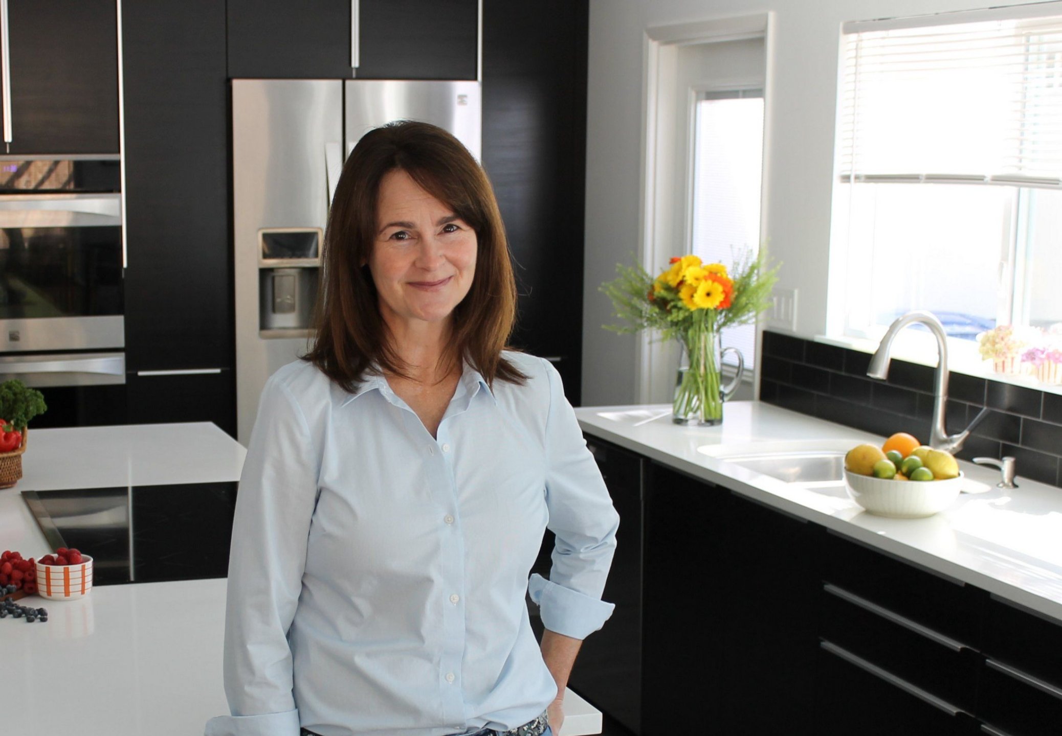 A woman with brown hair and a white shirt standing in a modern kitchen with black cabinets, a silver refrigerator, a white counter, a bowl of lemons and apples, a vase of yellow and orange flowers, and sunlight coming through a window.