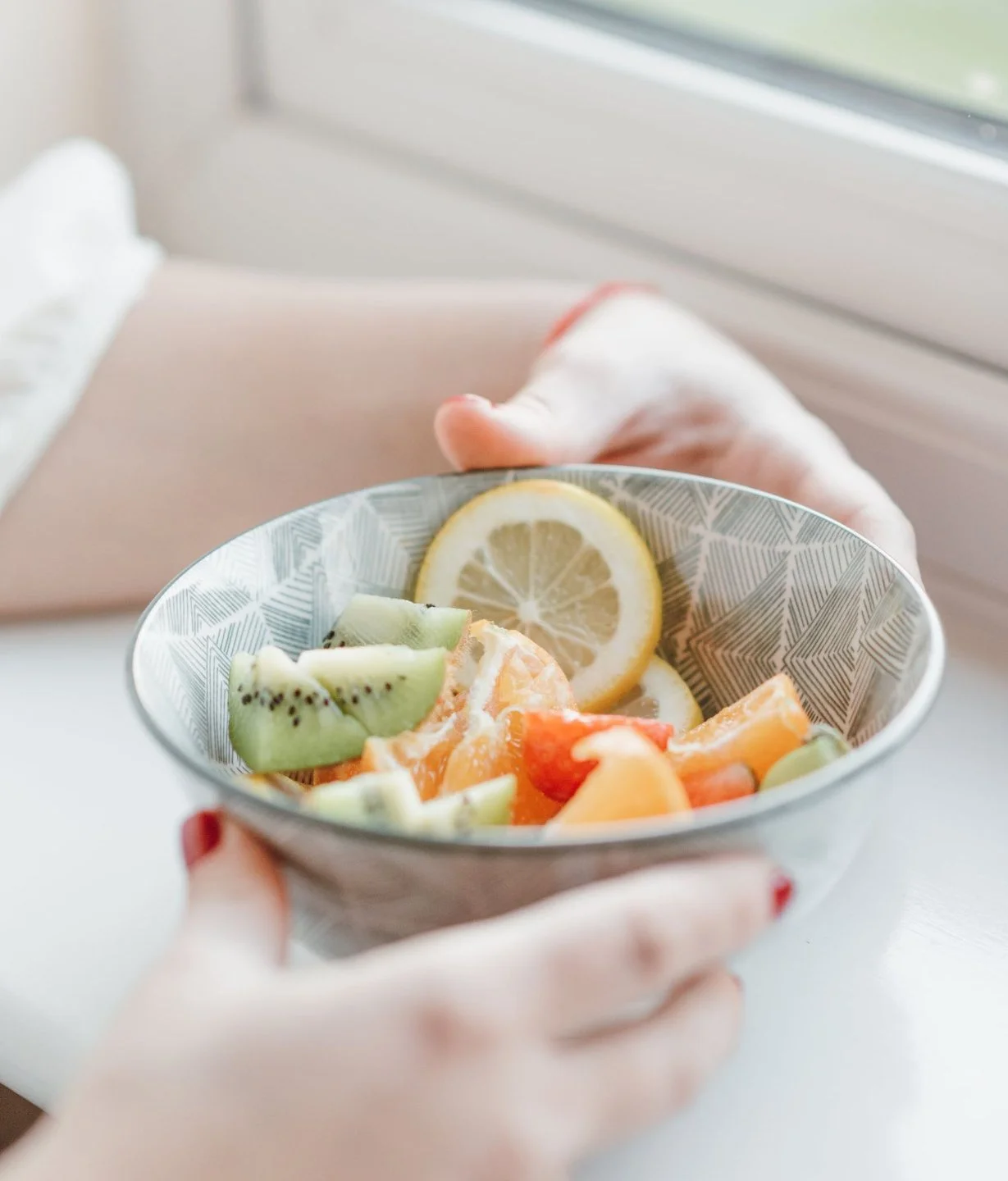 Close-up of hands holding a bowl of assorted cut fruits with lemon slices, near a window.