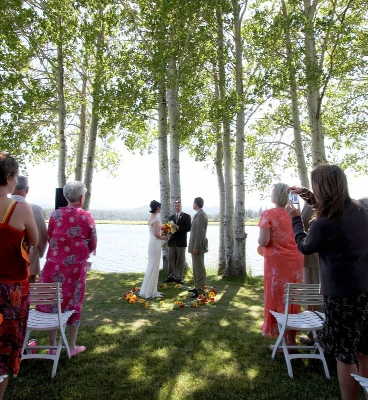 A wedding ceremony outdoors by a lake with trees, with the bride and groom exchanging vows, surrounded by guests taking photos.