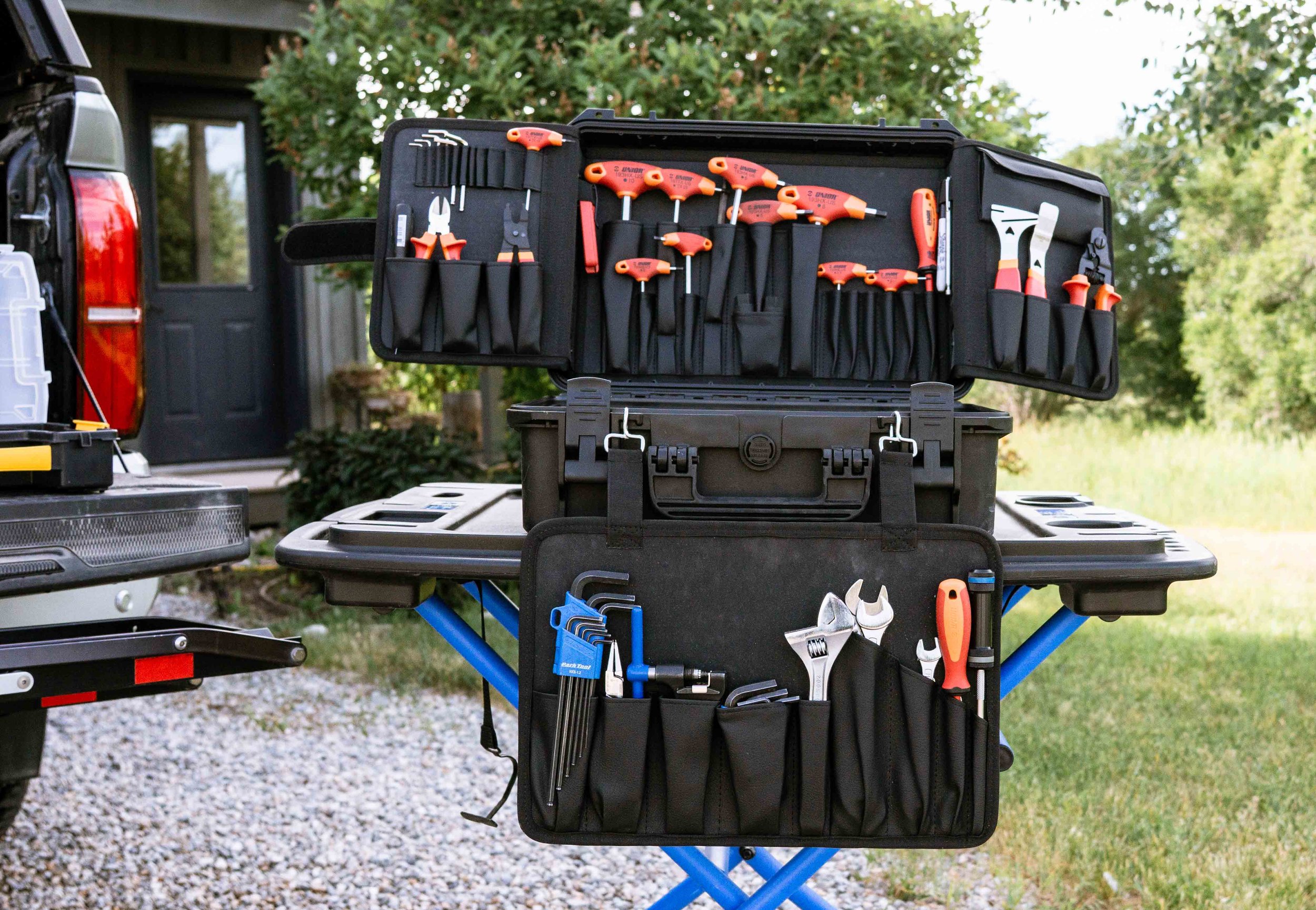 A black tool bag containing various orange-handled hand tools such as screwdrivers, wrenches, and pliers, set on a portable table outdoors with a background of green trees and a building.