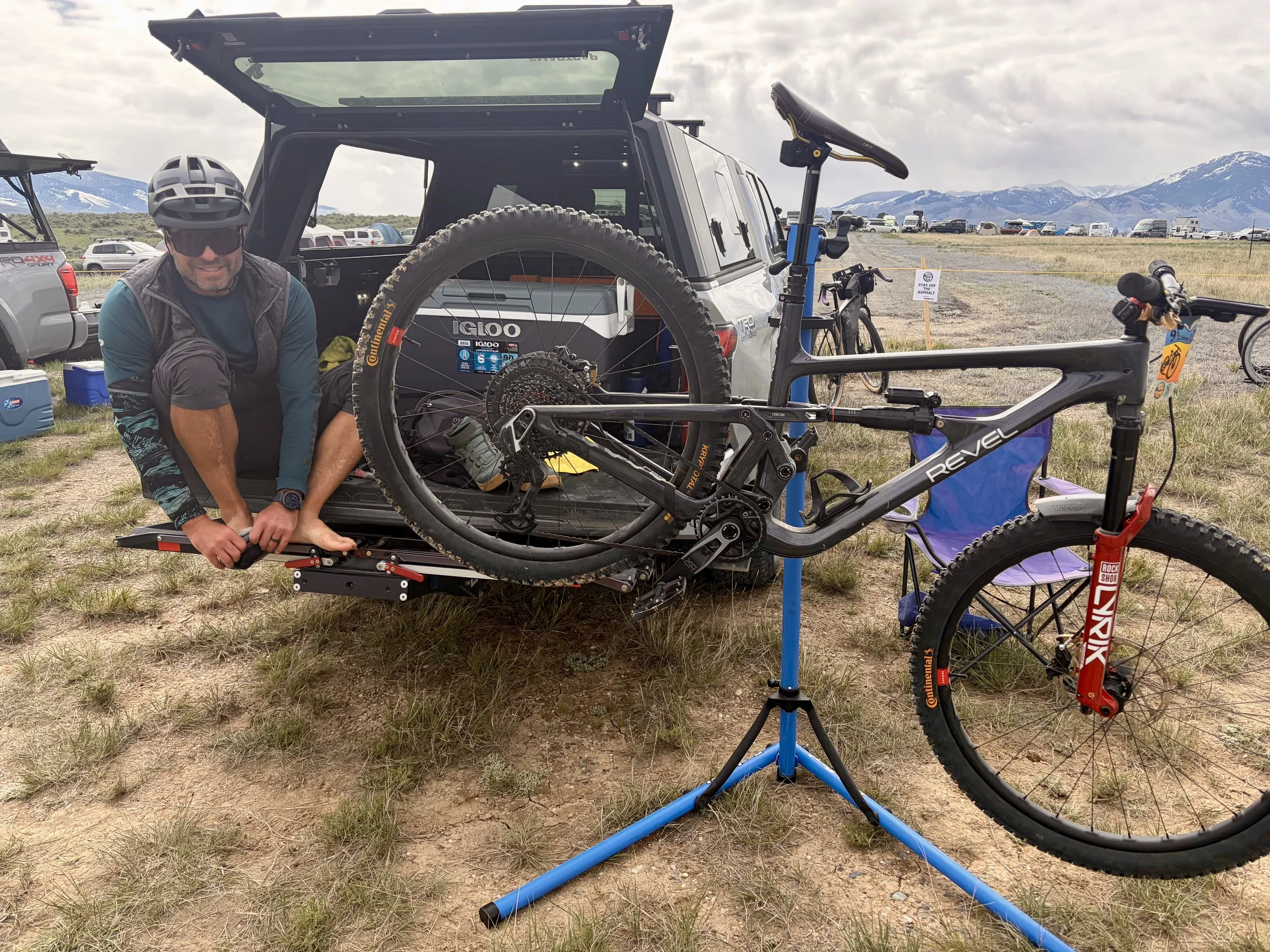 A man in a helmet crouches next to a mountain bike on a bike repair stand. The scene is outdoors in a field with mountains and cloudy sky in the background, surrounded by parked cars and tents.