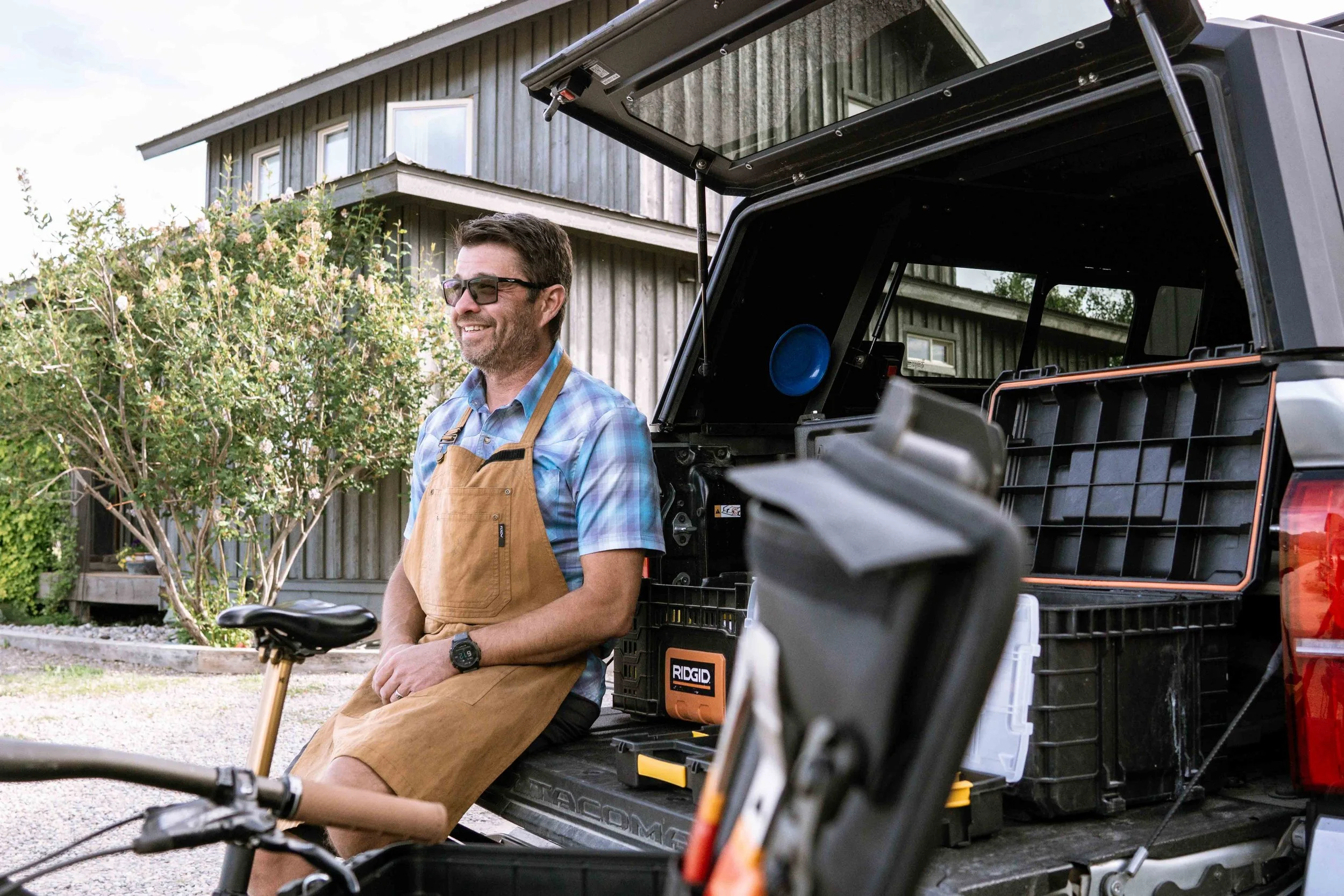 A man wearing sunglasses, a plaid shirt, and a brown apron sitting on a pickup truck tailgate. He is outdoors in front of a house with a gray wooden exterior, surrounded by greenery. There are tools and black storage boxes in the truck bed.