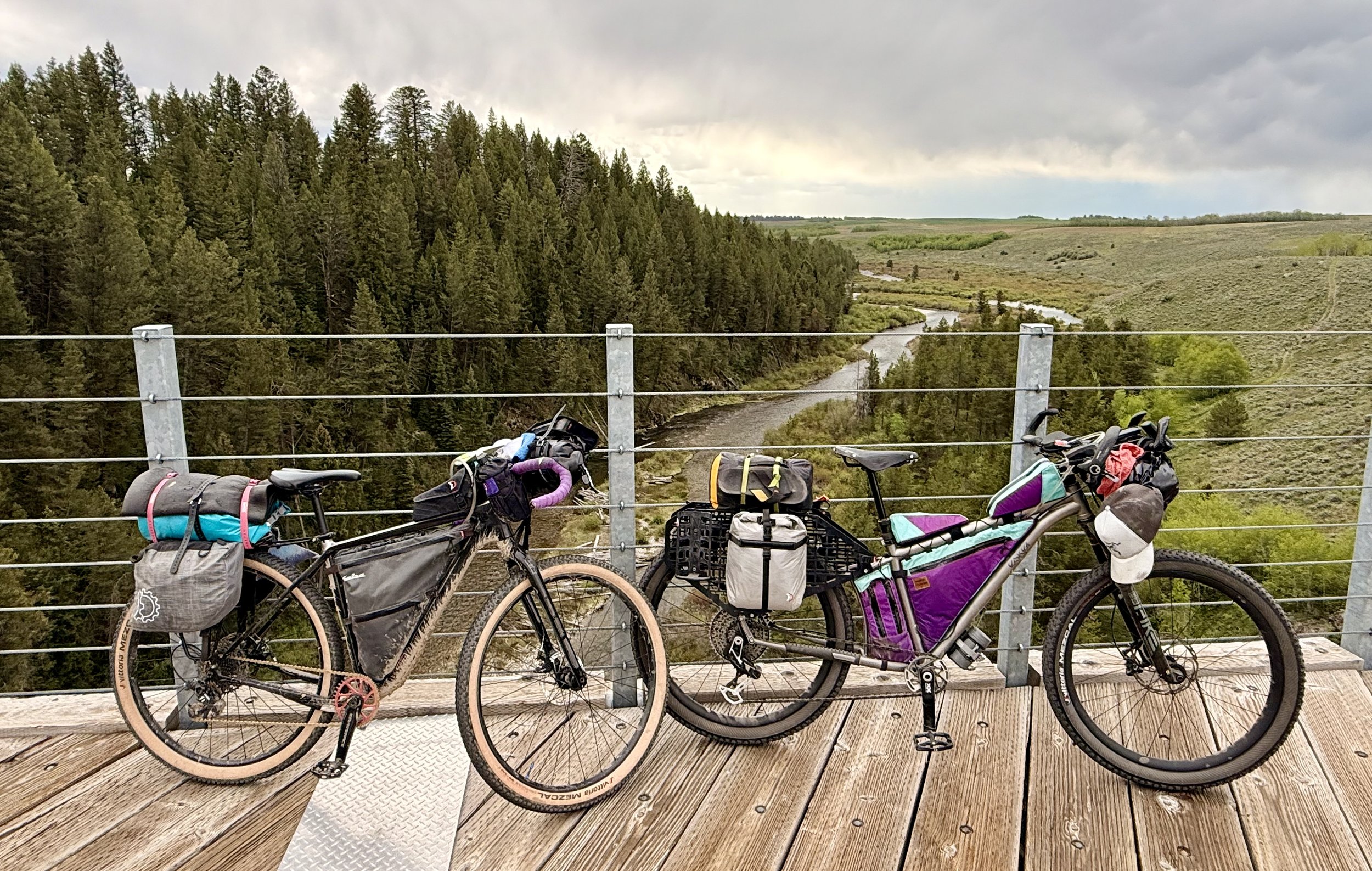 Two bicycles with gear parked on a wooden bridge overlooking a river and forested landscape under cloudy sky.