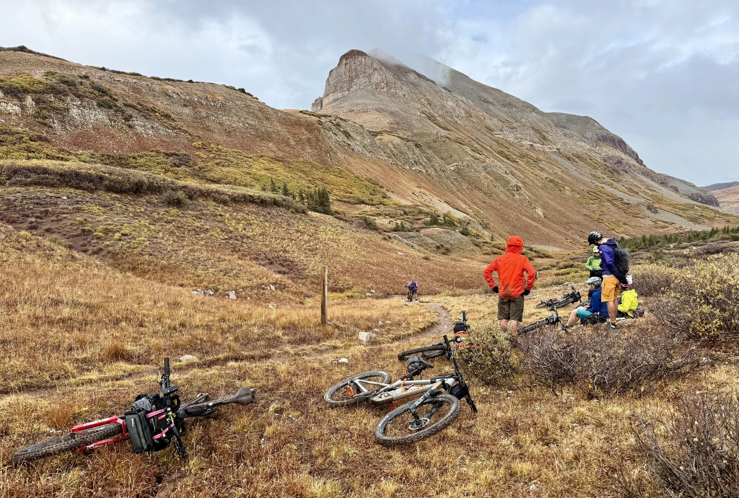 A group of mountain bikers resting on a trail in a mountainous landscape with a large rocky peak in the background. Bicycles are lying on the ground, and some bikers are sitting or standing, wearing helmets and outdoor gear.