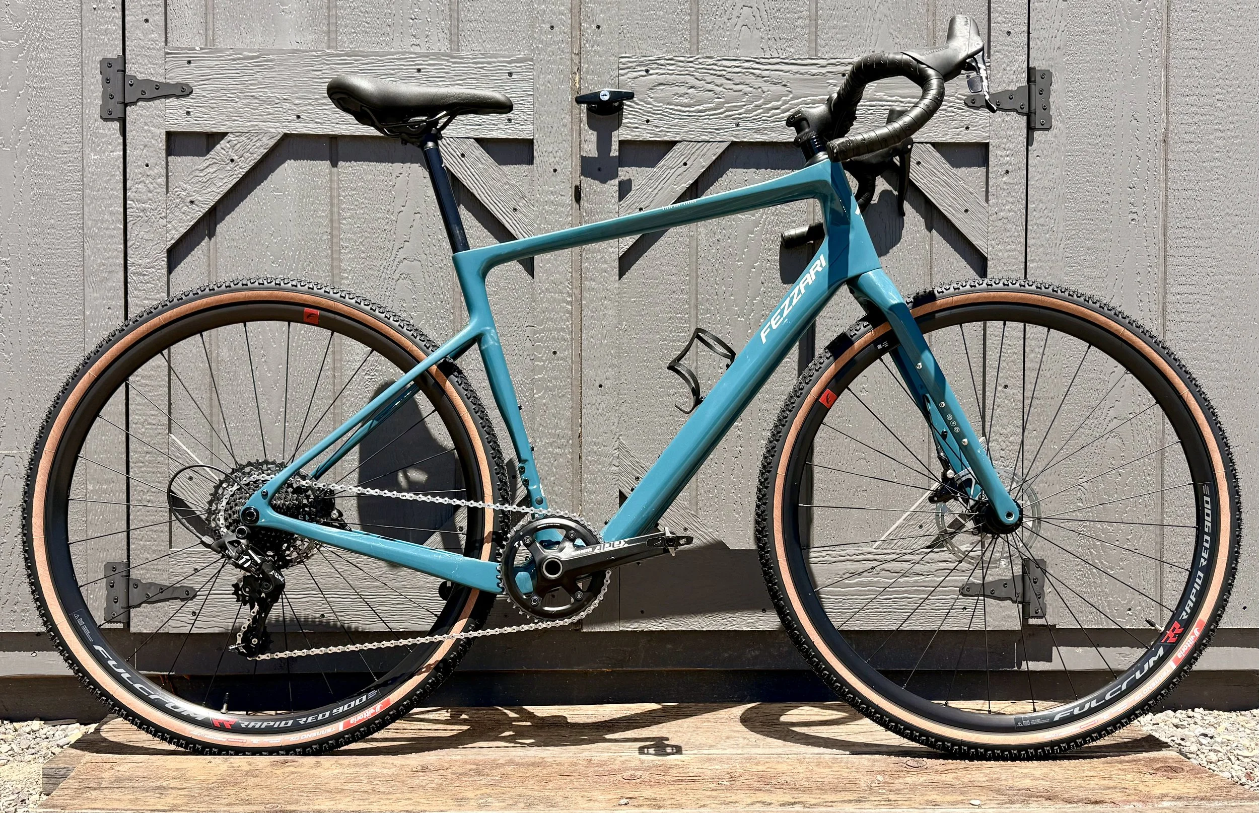 A blue racing bicycle with tan tires, positioned against a gray wooden shed door.