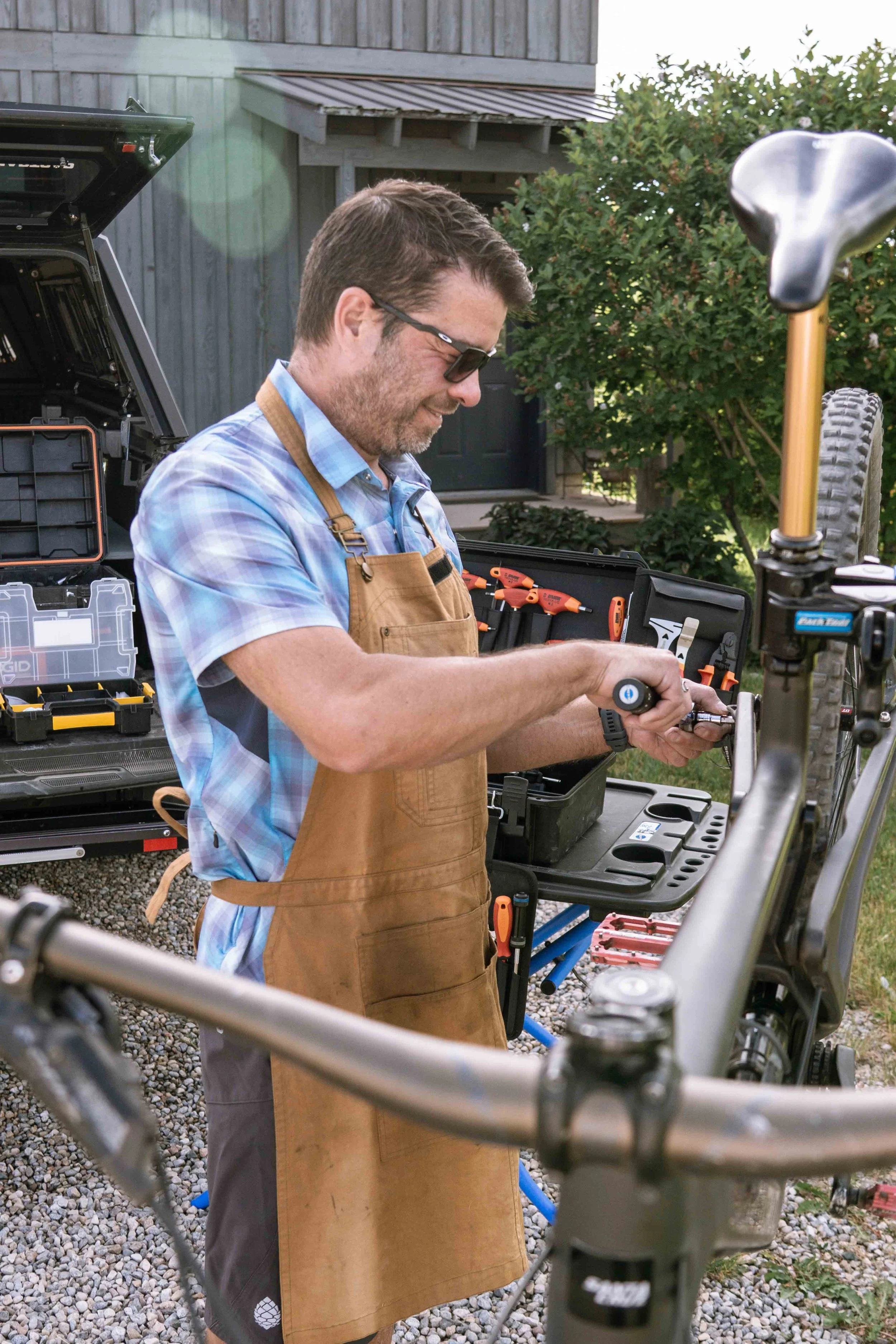 A man wearing sunglasses and a plaid shirt working on a bicycle, with a brown apron over his shirt. He is smiling and using tools from a black toolbox, set outdoors with a shed and greenery in the background.