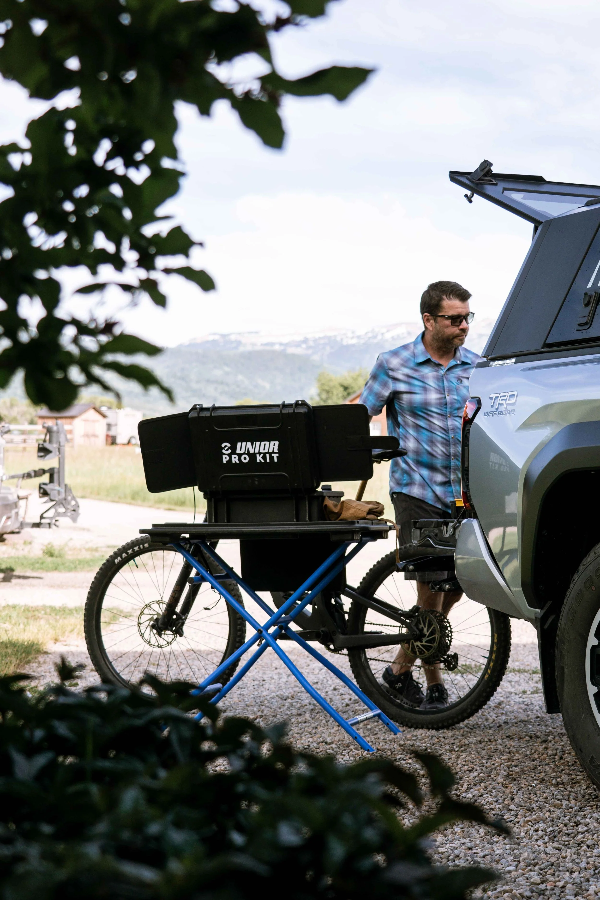 Man in sunglasses and plaid shirt working on a pickup truck, with a mountain landscape in the background, a bicycle with a Pro Kit toolbox nearby, and partial bushes in the foreground.