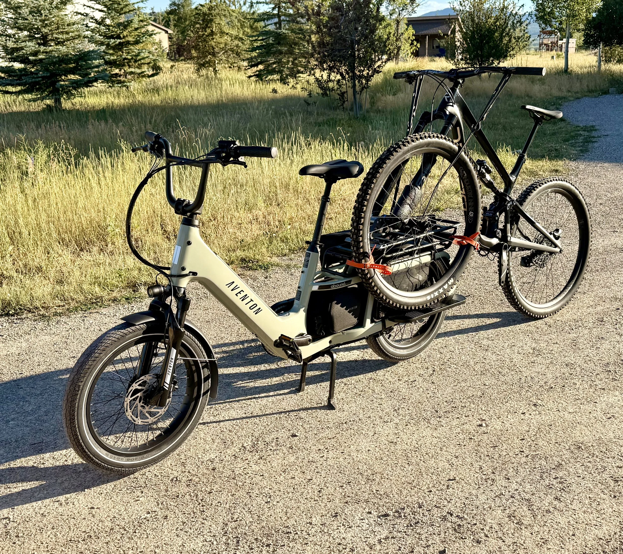 Two bicycles parked on a gravel path in a rural setting with trees and houses in the background, during daytime.