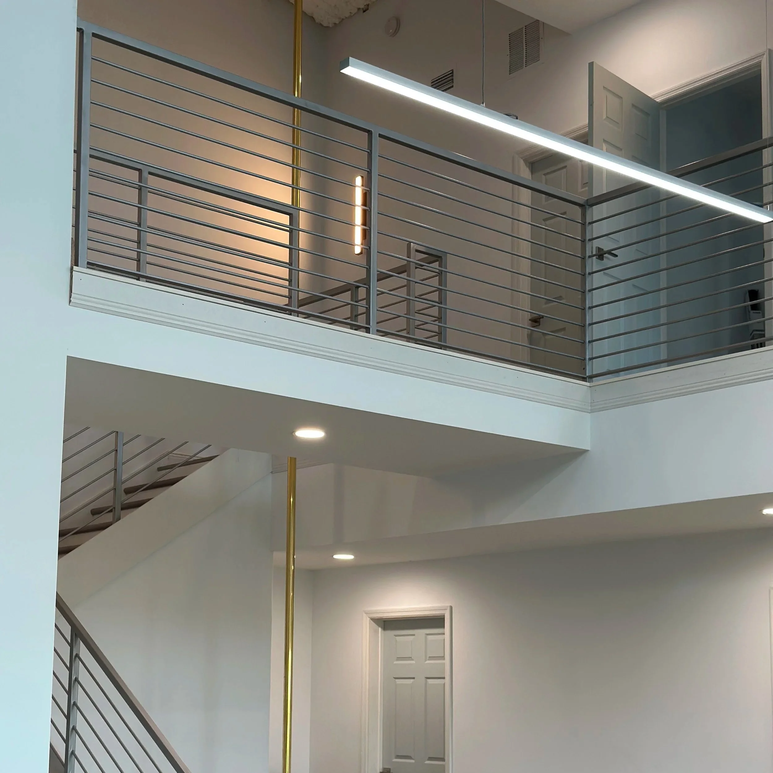 Interior view of a modern multi-story building, showing a railing on an upper floor with a sleek, horizontal design, a hanging linear light fixture, and partially open doors to rooms on the upper level. The ceiling has some exposed damage, and there are circular recessed lights on the ceiling.