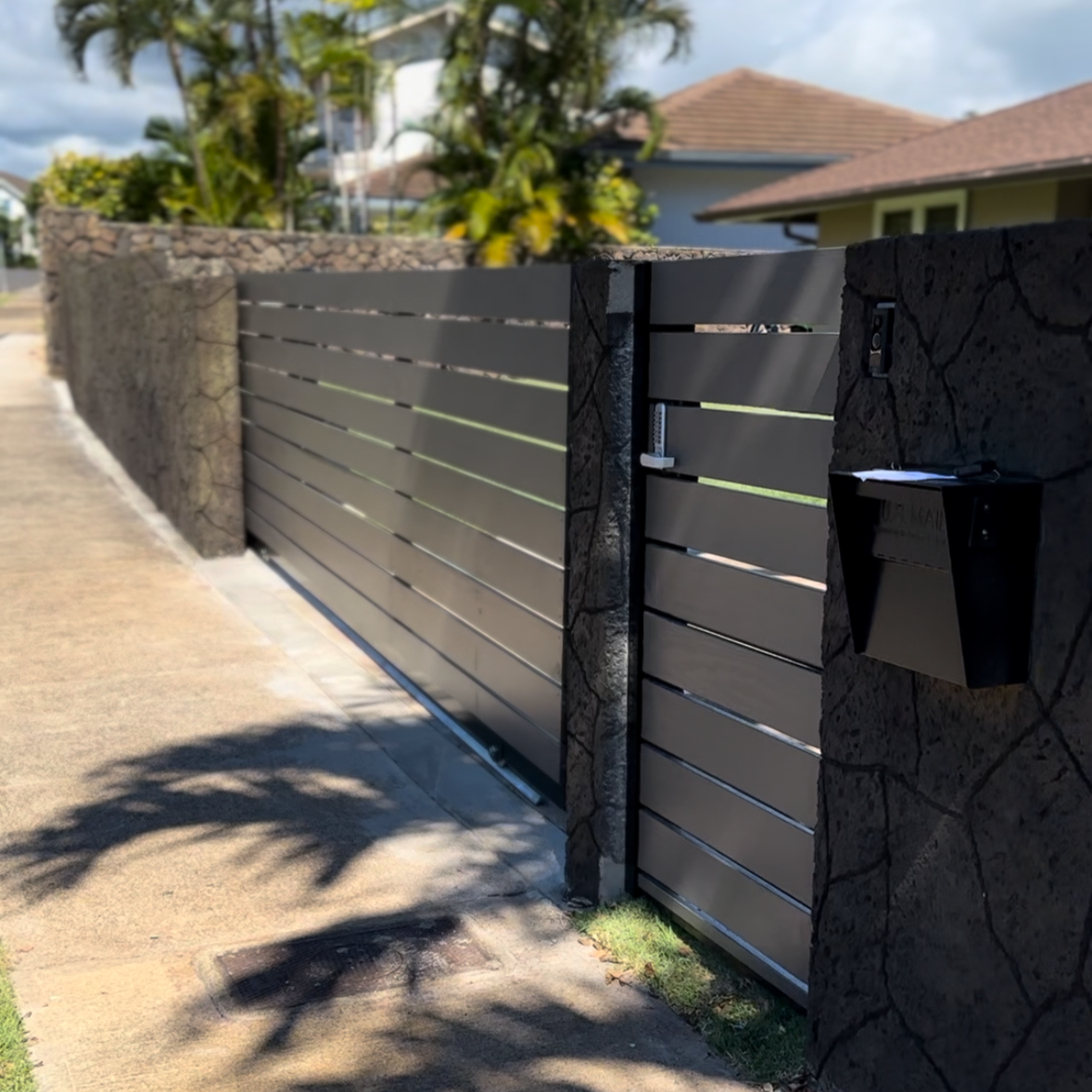 Modern sliding gate made of horizontal dark gray metal slats, flanked by stone pillars, with a mailbox and mail slot attached to the right pillar.