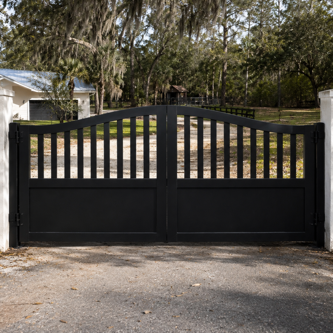 Black double driveway gates in front of a gravel driveway leading to a house and an outdoor area with trees and a wooden structure in the background.