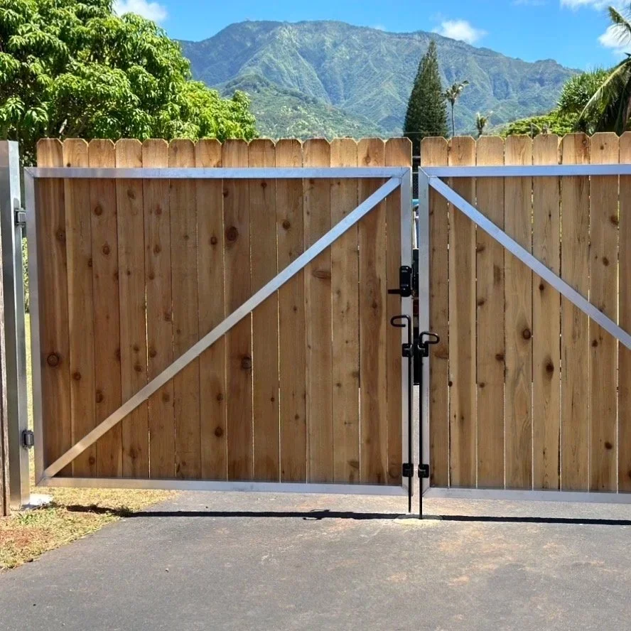 A wooden gate with metal frame and latch, set against a background of lush green trees and mountain landscape under a clear blue sky.