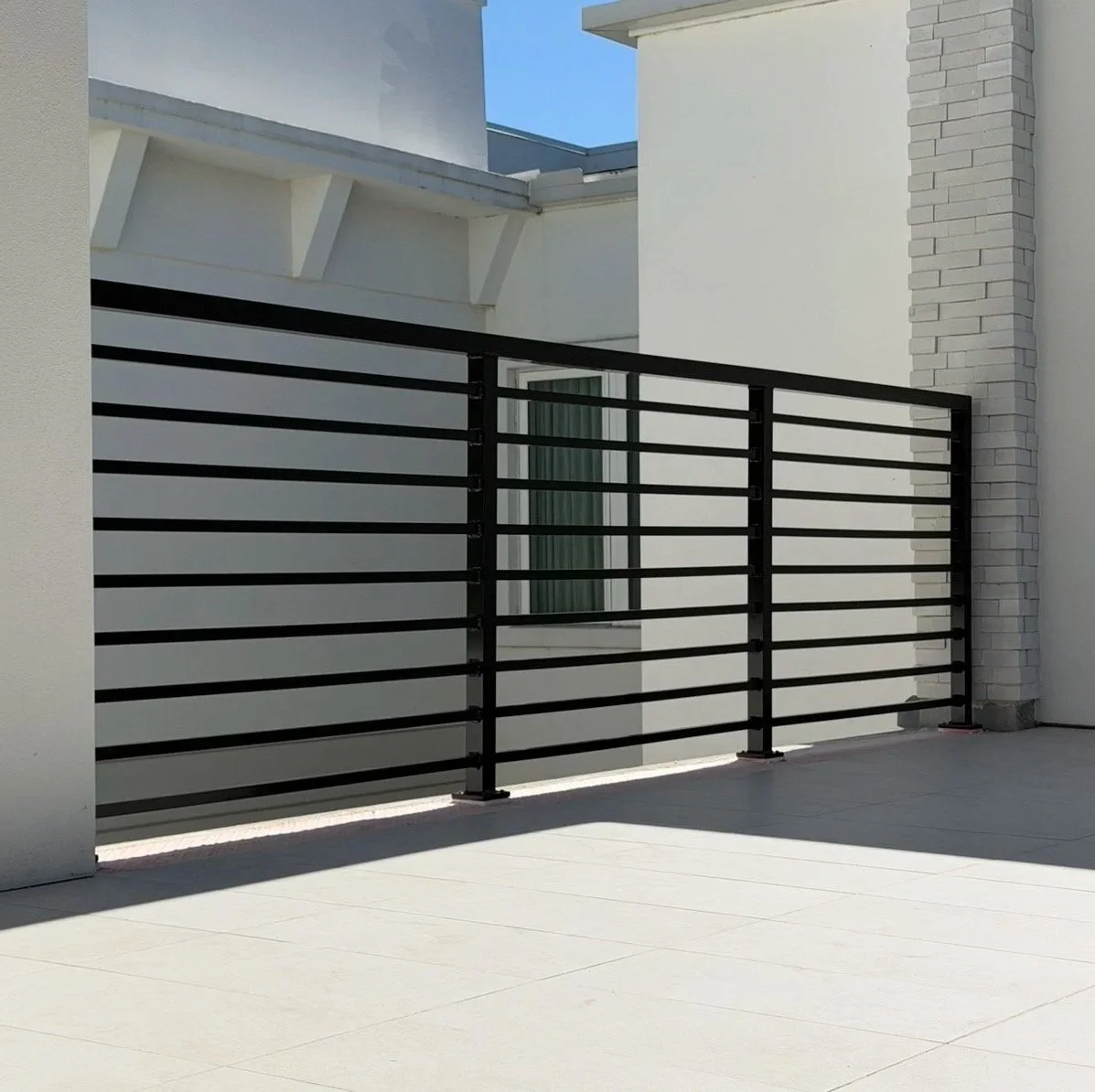 Modern balcony with black metal railing, white walls, and a brick column, with a clear blue sky in the background.