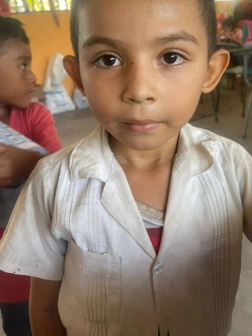 Close-up of a young boy with short dark hair and large eyes, wearing a white shirt, standing indoors with other children and classroom furniture in the background.