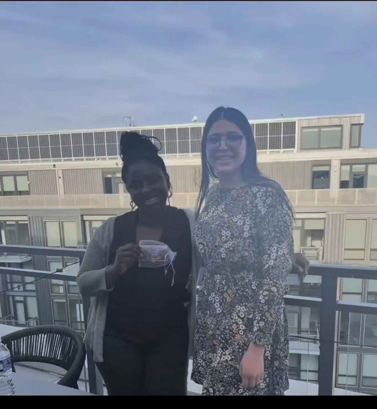 Two women standing on a balcony with buildings in the background. One woman is holding a face mask, and both are smiling.