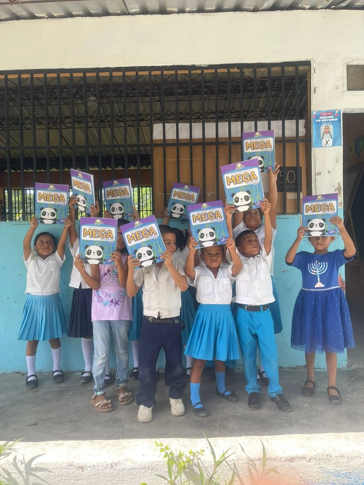Group of children in school uniforms holding up notebooks with panda and bamboo illustrations in front of a blue wall and black bars.