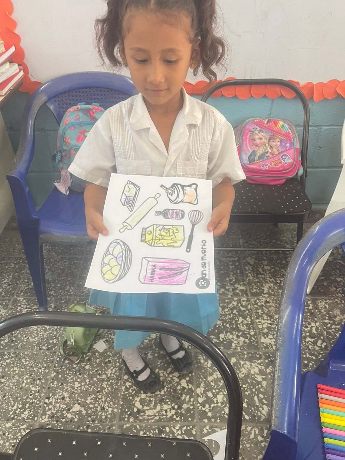 A young girl in a school uniform holding a piece of paper with colorful drawings of various food and drink items, standing in a classroom with chairs, backpacks, and a whiteboard in the background.