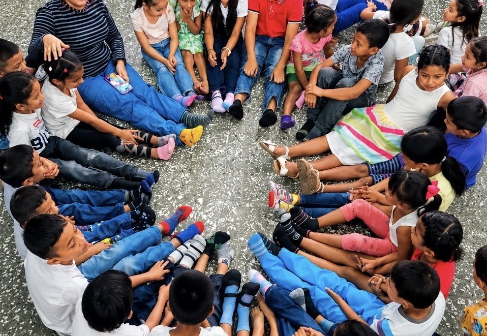 Group of children sitting in a circle on a speckled floor, engaging in a group activity, with a few adults present.