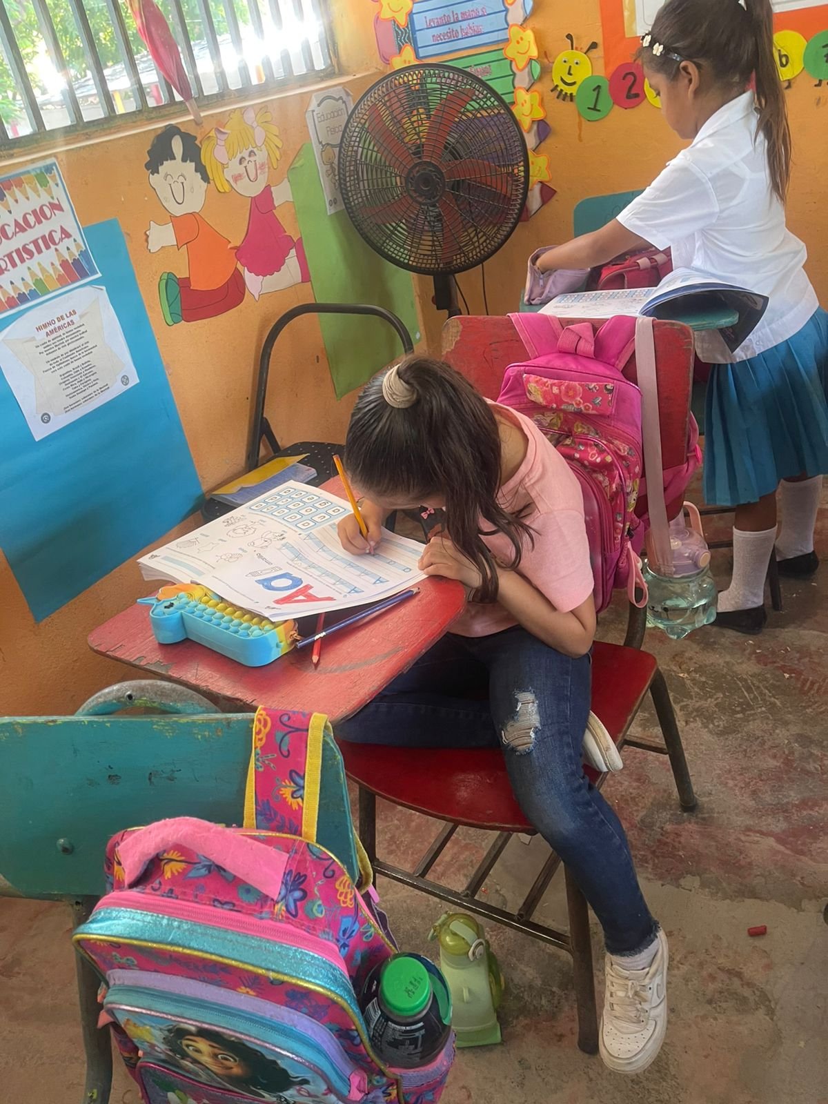 Two young girls in a classroom, one sitting at a desk coloring a worksheet and the other standing with a book and backpack. The classroom has colorful posters and murals on the wall, and backpacks hang on the chairs.