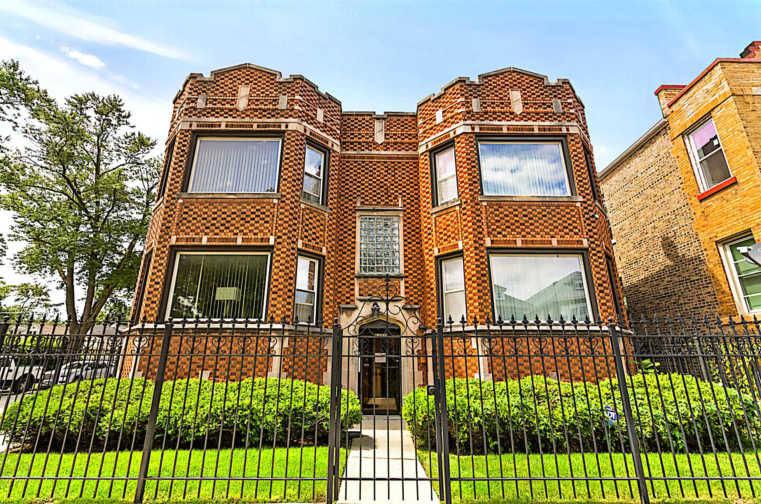 A brick building with an ornate design, metal gate, and large windows, surrounded by a lush green lawn and trees, under a blue sky.