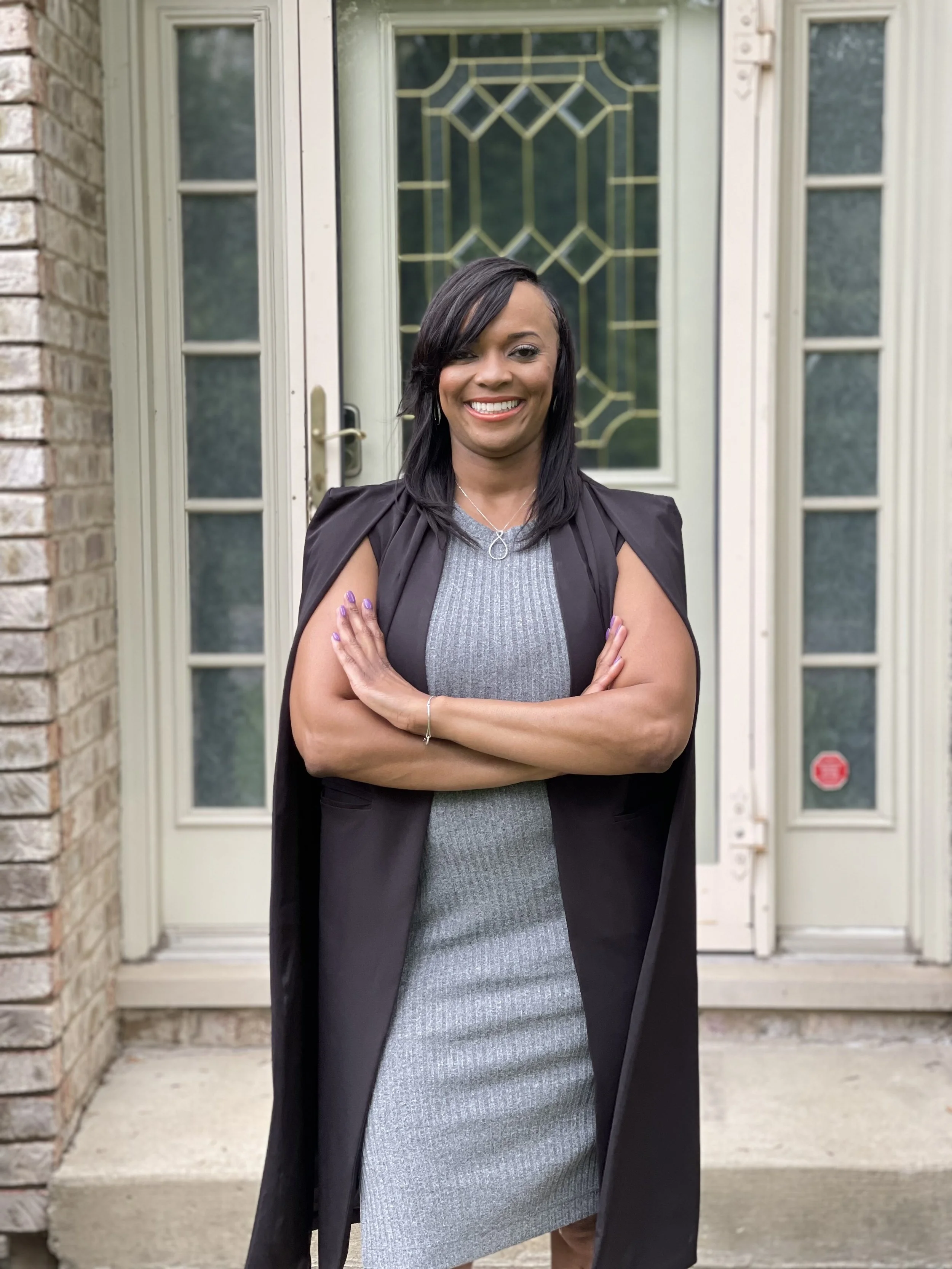 A smiling woman with black hair, wearing a gray dress and black blazer, standing arms crossed in front of a house door with glass and decorative gold design.