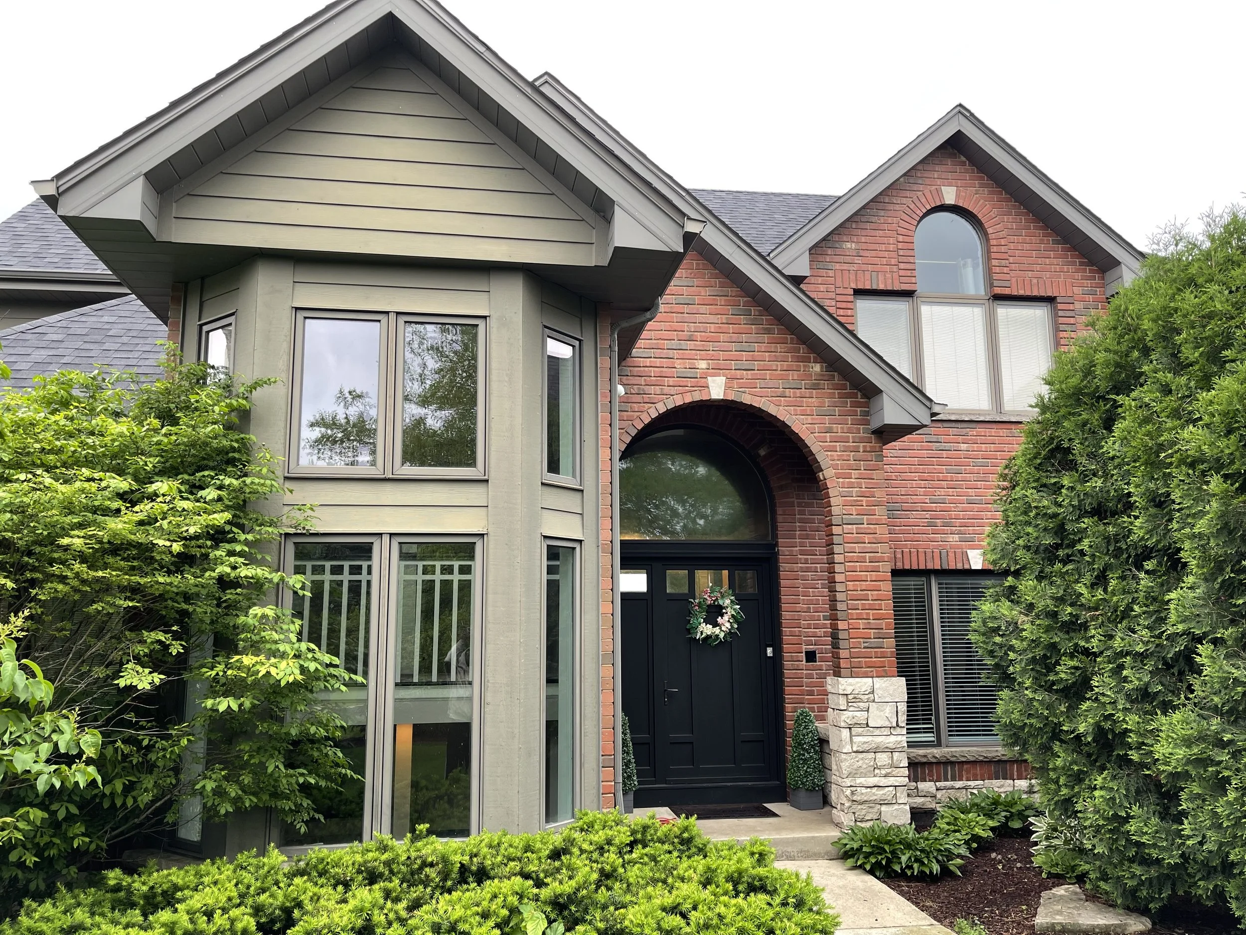 A modern house with a combination of grey siding and red brick, featuring large windows, a black front door with a holiday wreath, and lush green plants and trees in the front yard.