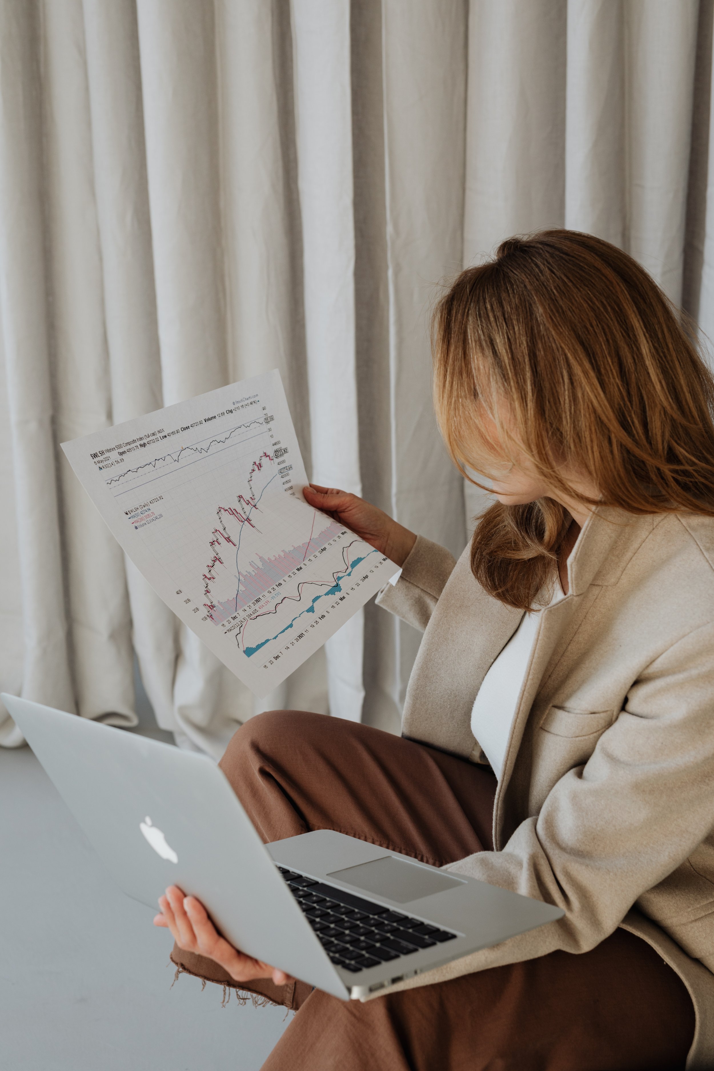 A woman with brown hair sits indoors, holding a printed stock market chart, a silver MacBook on her lap, and a laptop in her hand, with beige curtains in the background.