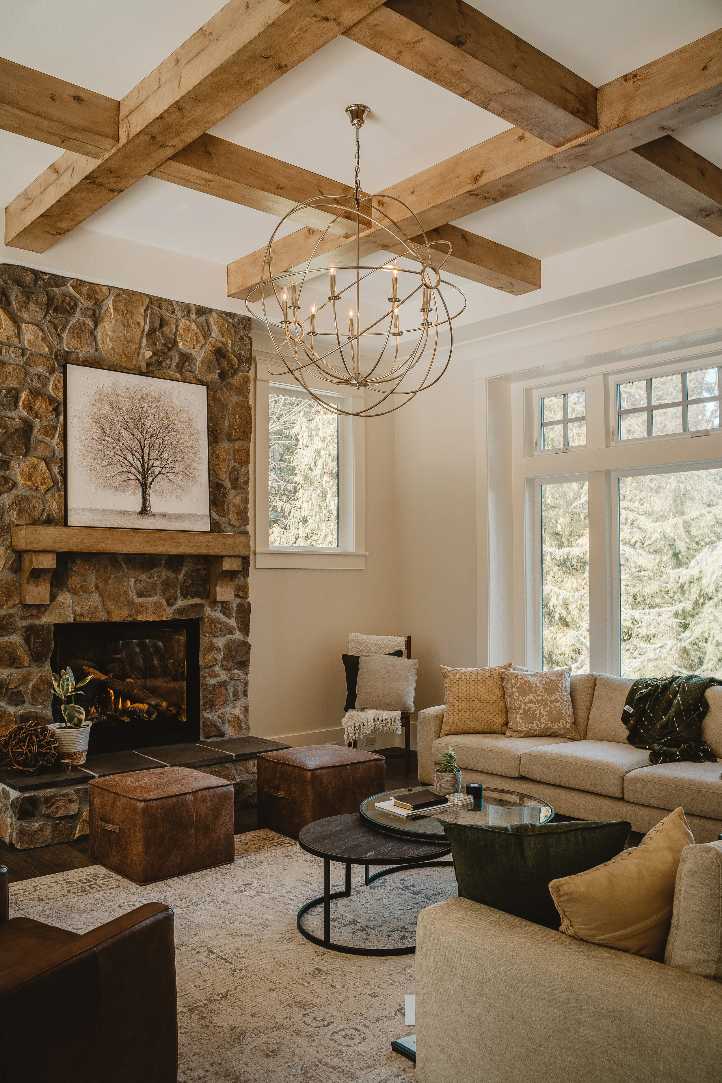 Living room with a stone fireplace, wooden beam ceiling, modern chandelier, large window showing trees, and beige sofa with pillows.