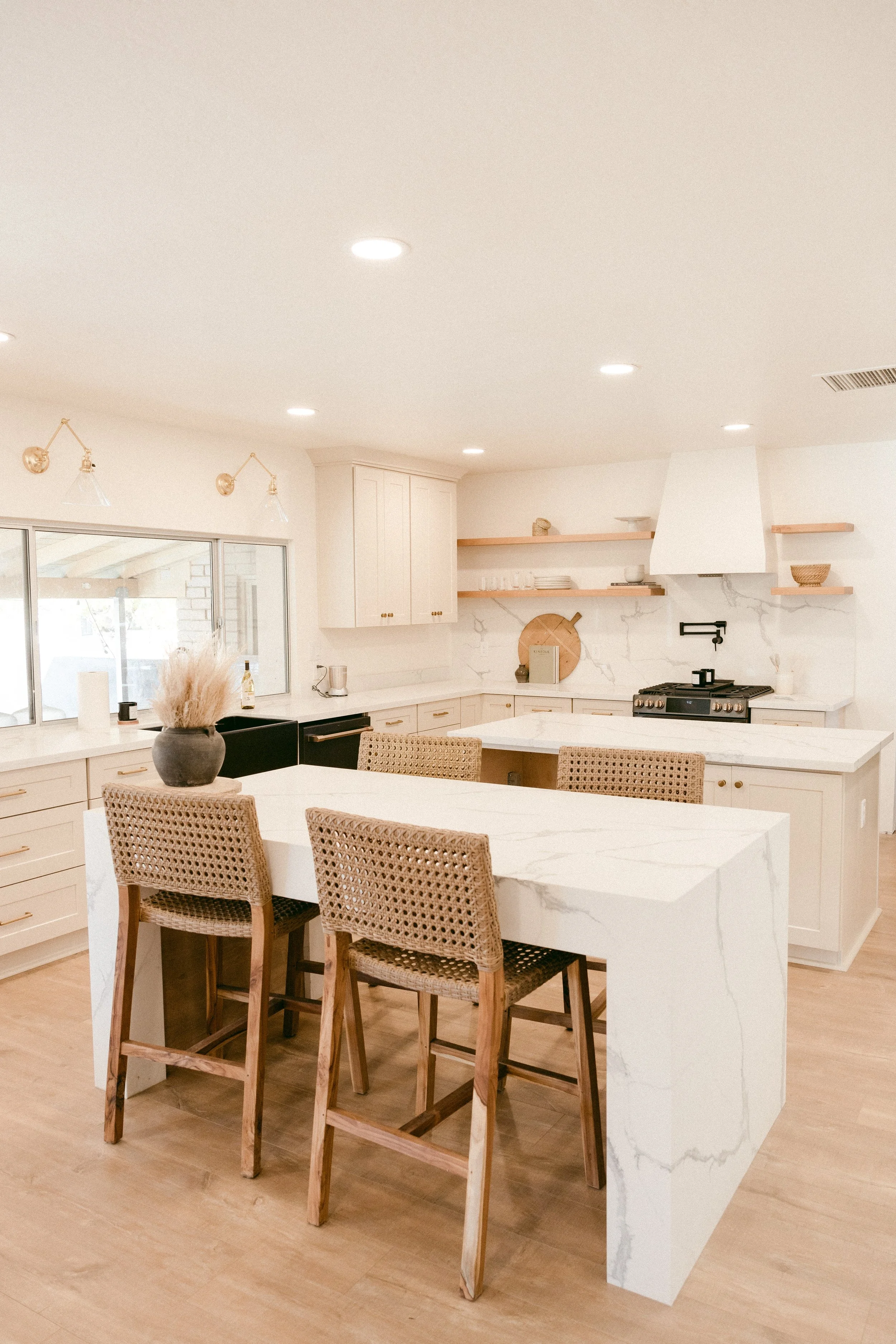 Modern kitchen with white marble island, four wicker barstools, white cabinetry, open wooden shelves, black stove, large window, and minimalist decor.