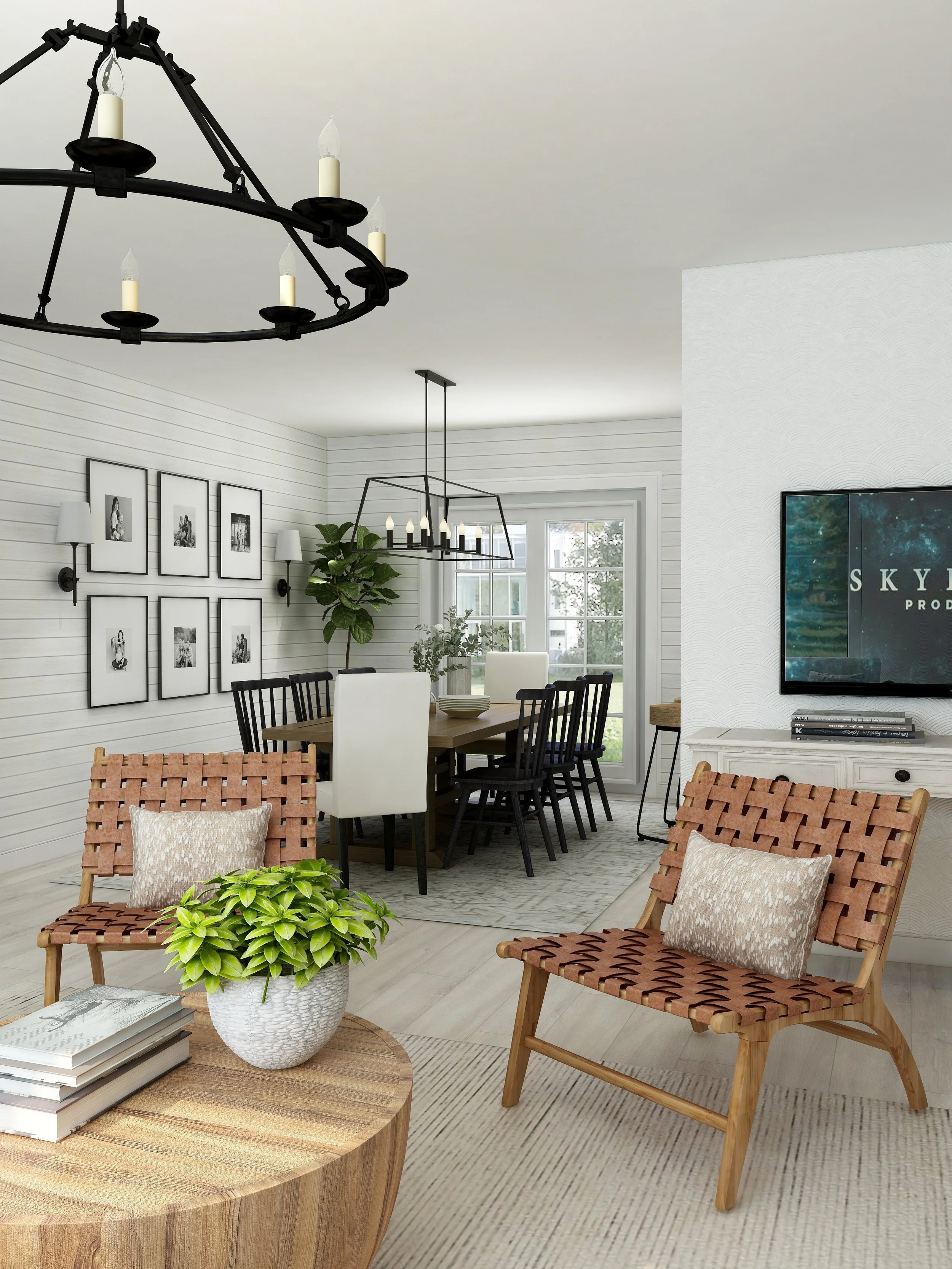 A living room or dining area with a wooden coffee table, potted plant, two woven chairs with pillows, and a dining table with black chairs and framed photographs on the white shiplap wall. There are two lighting fixtures, a modern chandelier and a chandelier with candles, hanging from the ceiling. A television is mounted on a textured wall to the right.