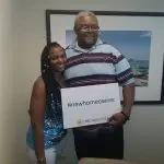 A smiling young woman and a smiling man holding a sign that says www.homeschooling.com, standing in front of a framed photo on a wall.
