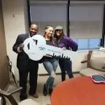 Three people standing indoors, one holding a large key prop, all smiling.