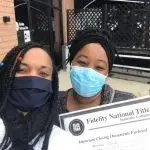 Two women wearing masks, holding a government document or certificate, possibly related to voting, in front of a black metal fence.