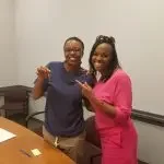 Two women standing together in an office, smiling and making peace signs with their hands.