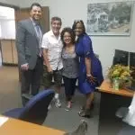 Four people standing together indoors, in an office setting with a painting and a plant on a table nearby.