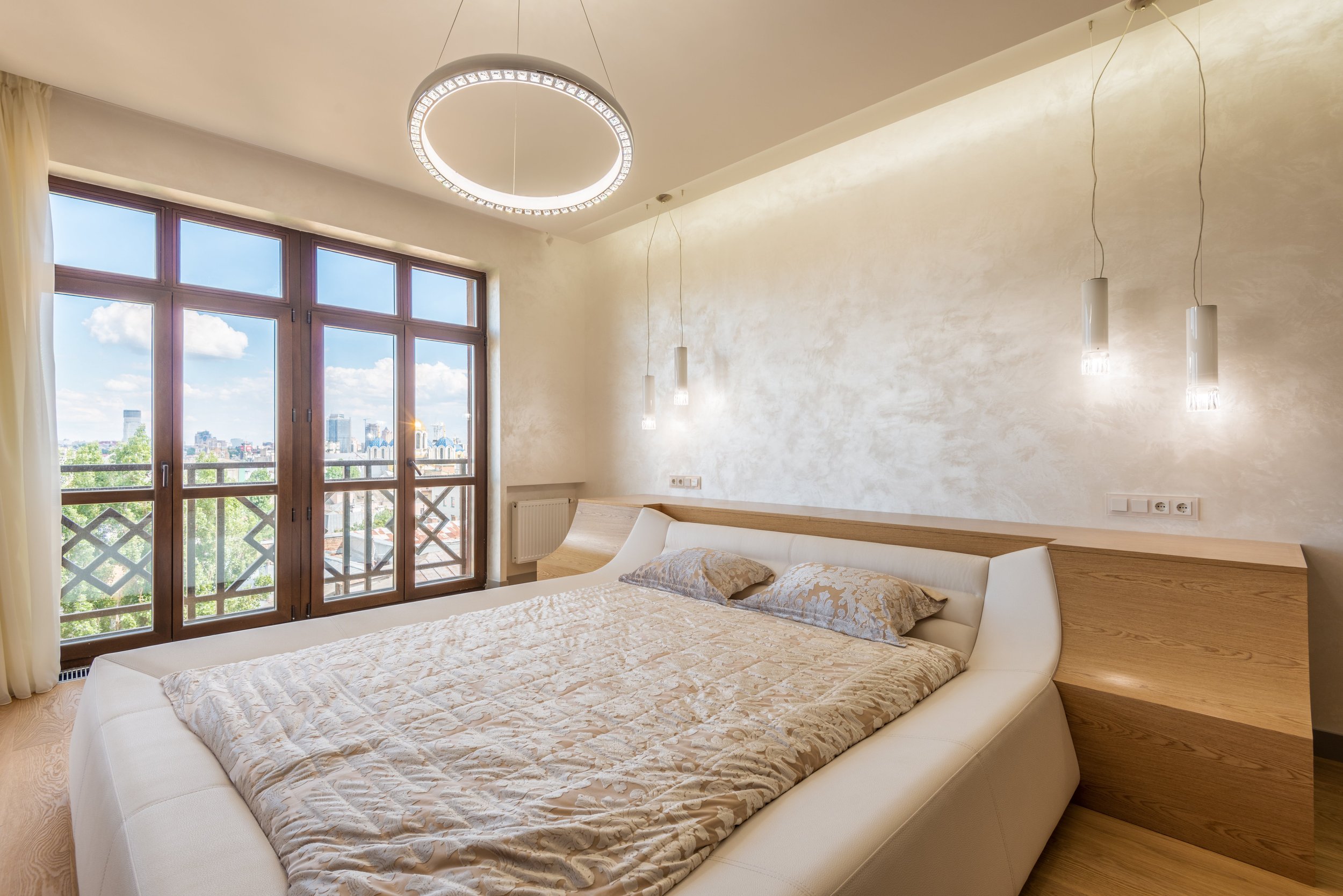 A bedroom with a large window and balcony doors, a white modern bed with a beige quilt and pillows, a wooden headboard, hanging ceiling lights, and cityscape view outside.