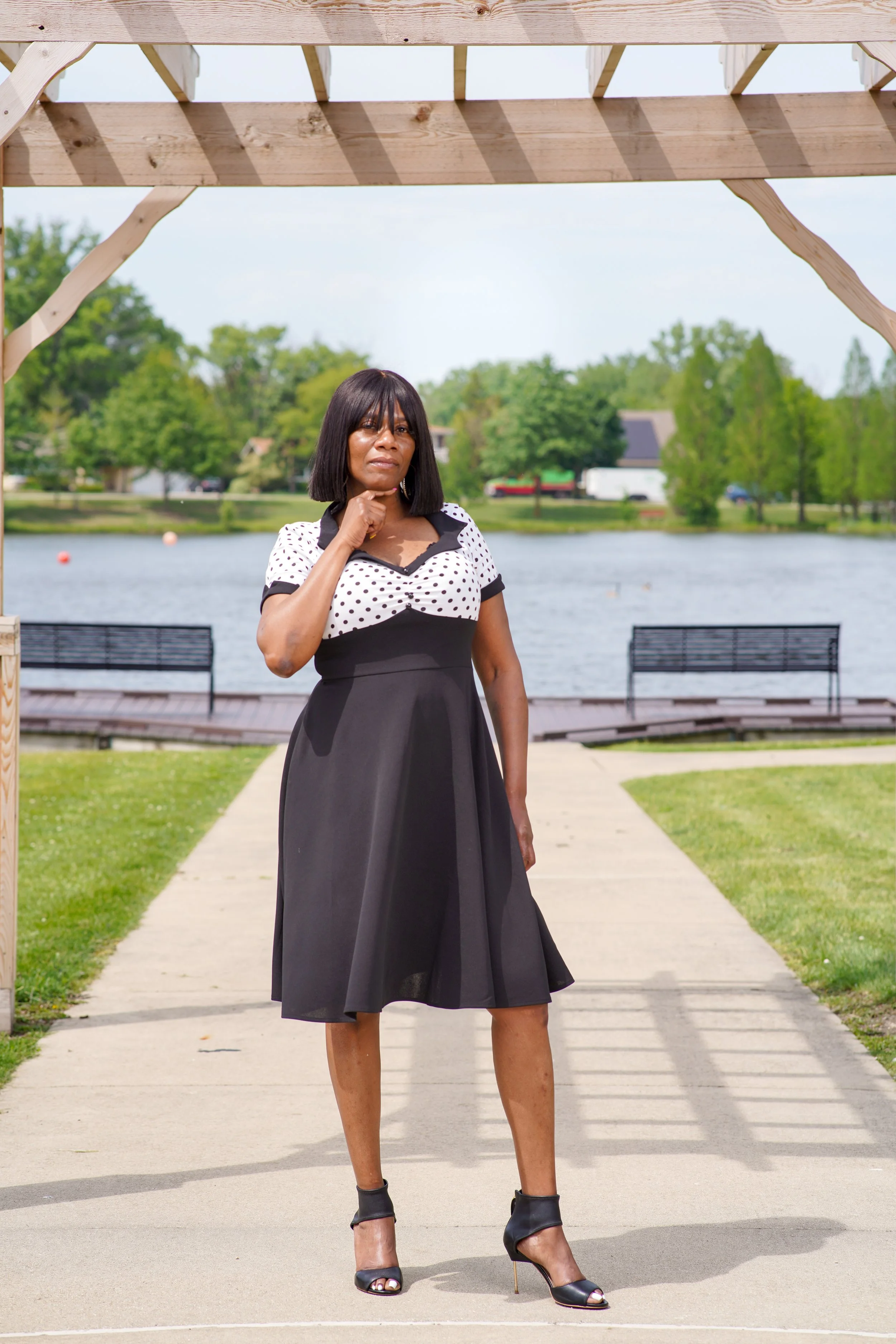 Woman standing outdoors under a wooden structure, near a pond with trees and houses in the background, wearing a black and white dress and heels.