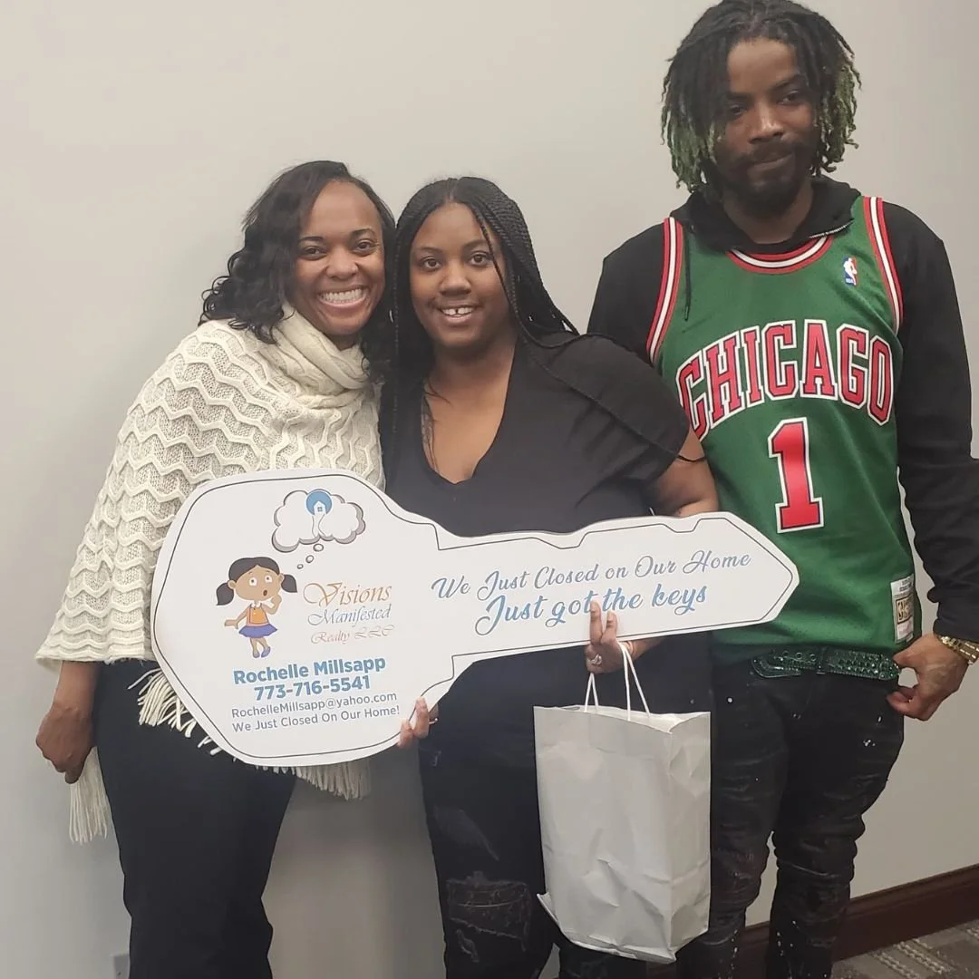 Three people standing together, holding a large speech bubble-shaped sign that reads "We Just Closed on Our Home. Just got the keys." The woman on the left is smiling, wearing a cream-colored shawl, the woman in the middle is also smiling, and the ma