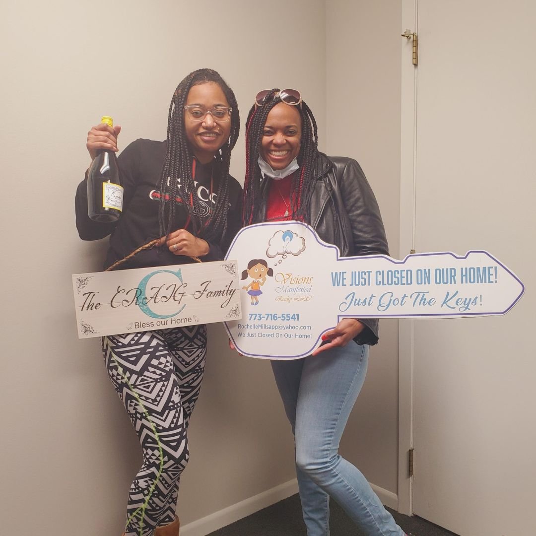 Two women smiling and holding signs celebrating closing on a home. One woman holds a small bottle of champagne, and the other holds a sign that says 'WE JUST CLOSED ON OUR HOME! Just Got The Keys!' The woman on the left has glasses and braided hair, 