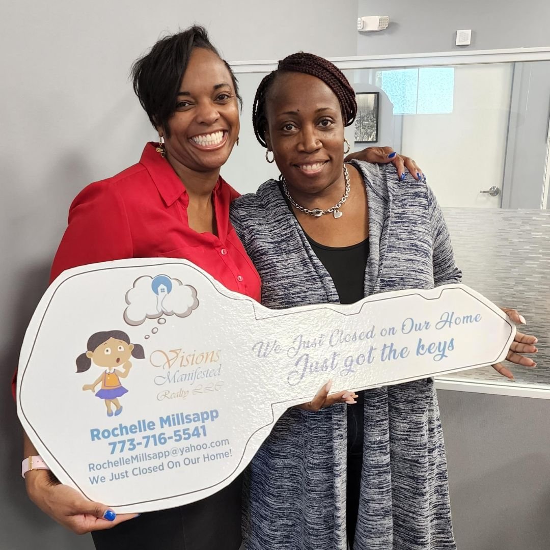 Two women smiling and holding a large key-shaped sign that reads 'We Just Closed on Our Home, Just got the keys.' One woman is in a red shirt, and the other is in a gray cardigan. The sign displays contact information for Rochelle Millsap of Visions 