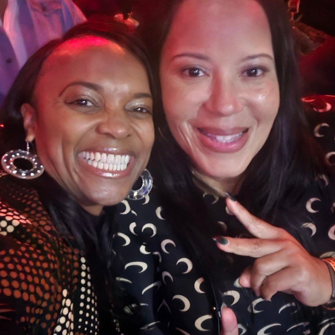 Two women taking a close-up selfie, smiling, one making a peace sign, wearing black shirts with white moon patterns, and large earrings.
