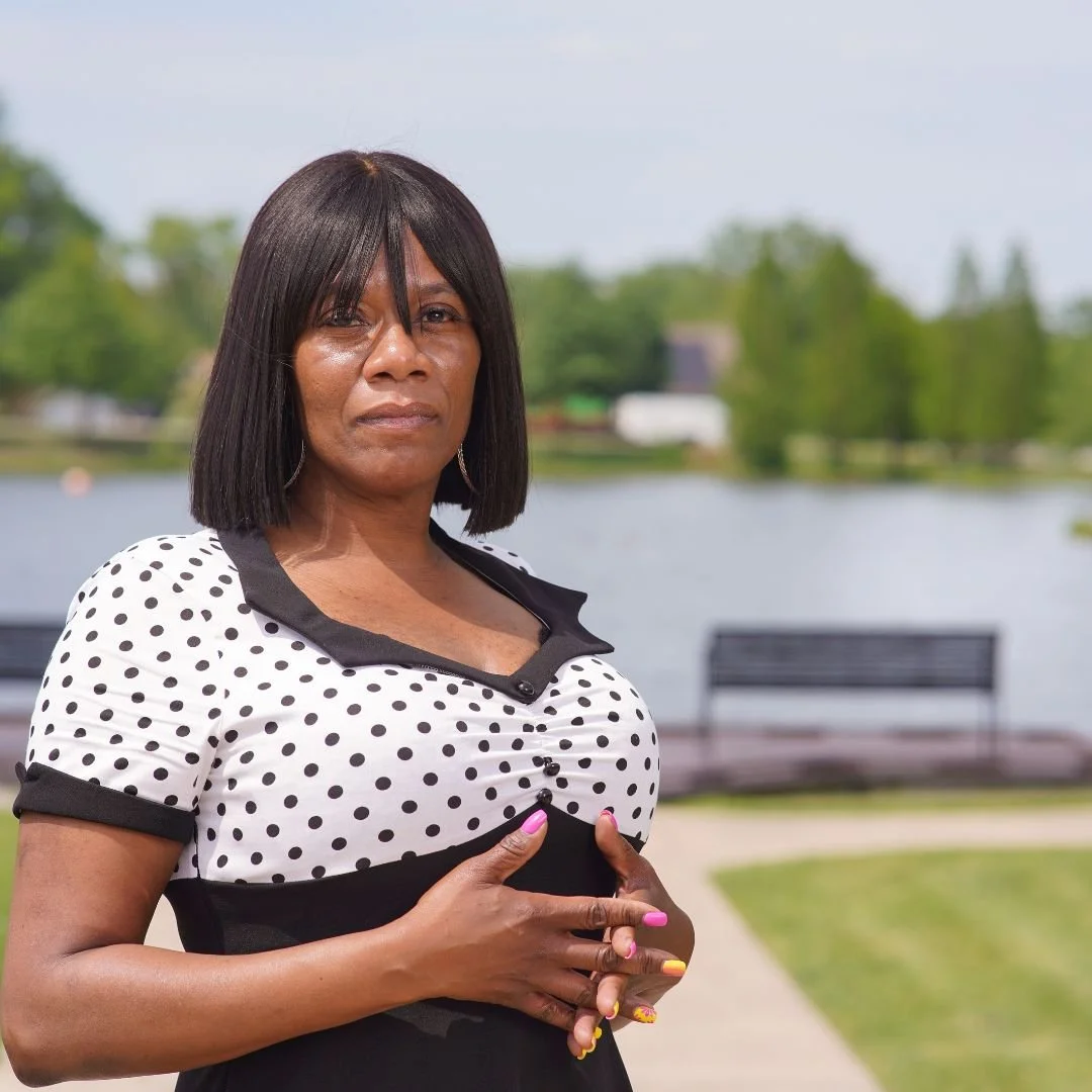 A woman with shoulder-length black hair and dark skin, wearing a white blouse with black polka dots and black trim, stands outdoors near a body of water with green trees and benches in the background.