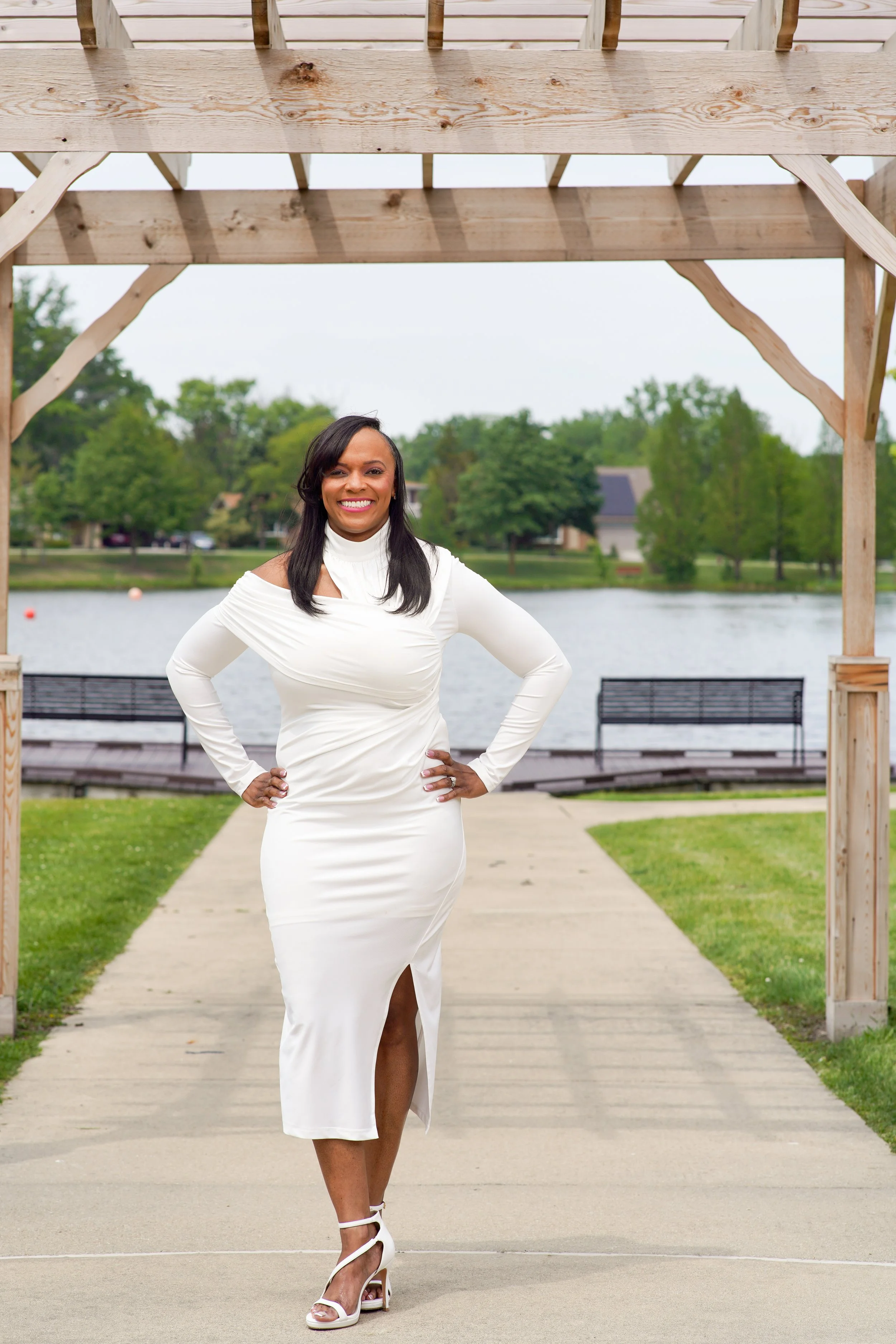 A woman in a white dress and high heels standing outdoors on a sidewalk by a lake, with a wooden arch above her, surrounded by greenery and houses in the background.