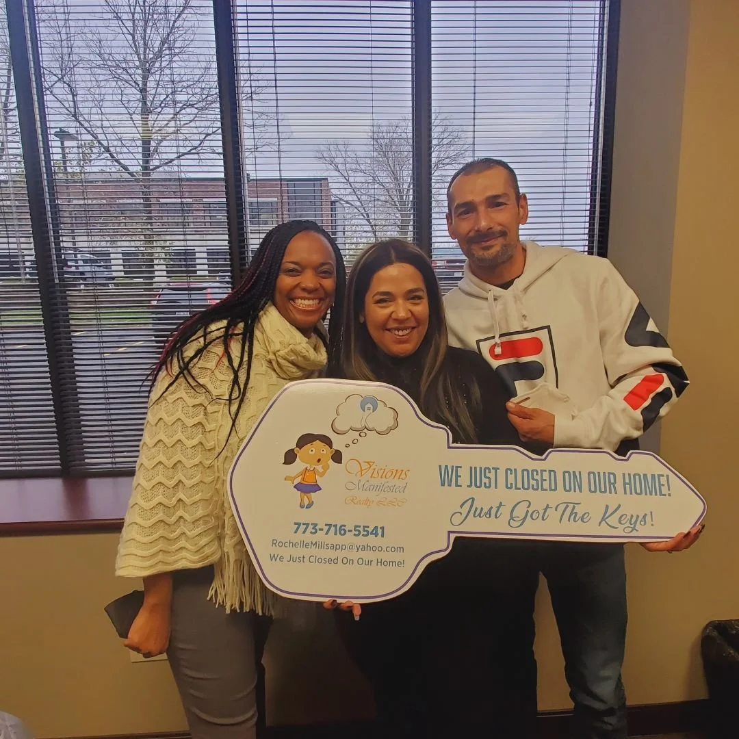 Three smiling people holding a sign that says 'We just closed on our home! Just Got The Keys!' inside a room with a large window and blinds, with leafless trees outside.
