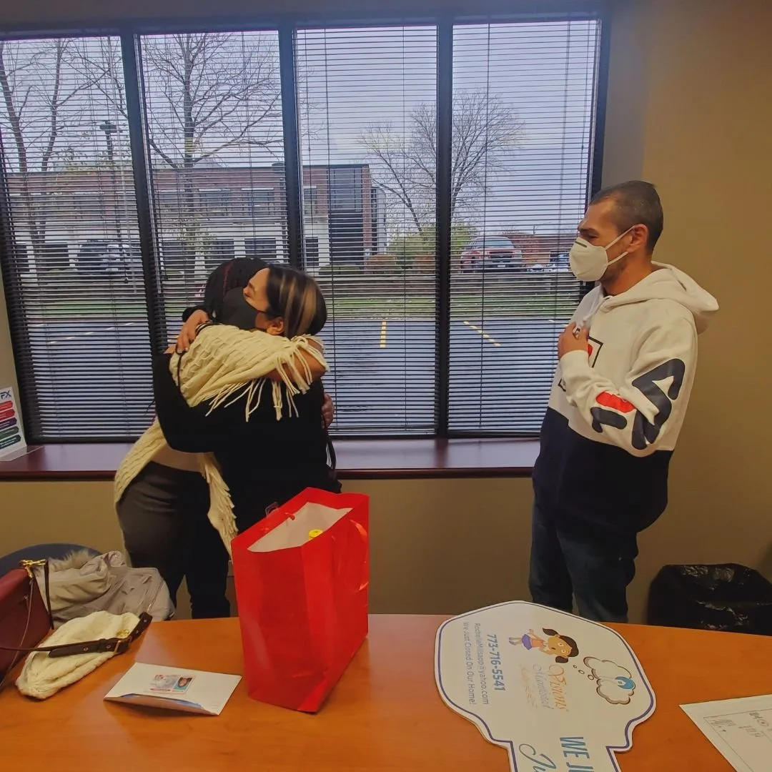 Two women hugging each other inside, a man standing near them, all wearing face masks. The women are embracing warmly near a window with blinds, and the table in front has a red gift bag, some papers, and belongings.