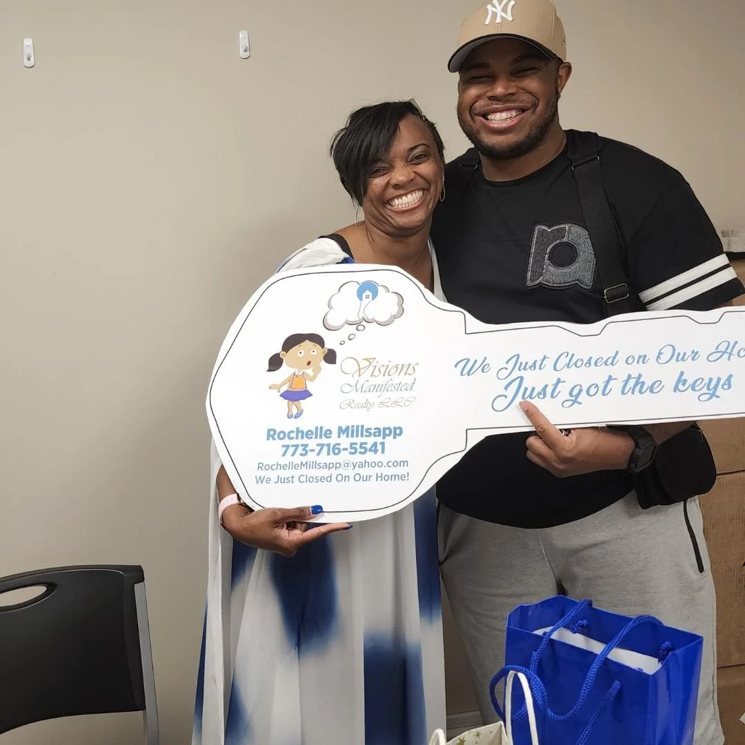 Two smiling people standing together, holding a large key-shaped sign that says, 'We Just Closed on Our Home. Just got the keys.' The woman is wearing a white and blue tie-dye dress, and the man is wearing a black t-shirt, beige cap, and gray sweatpa