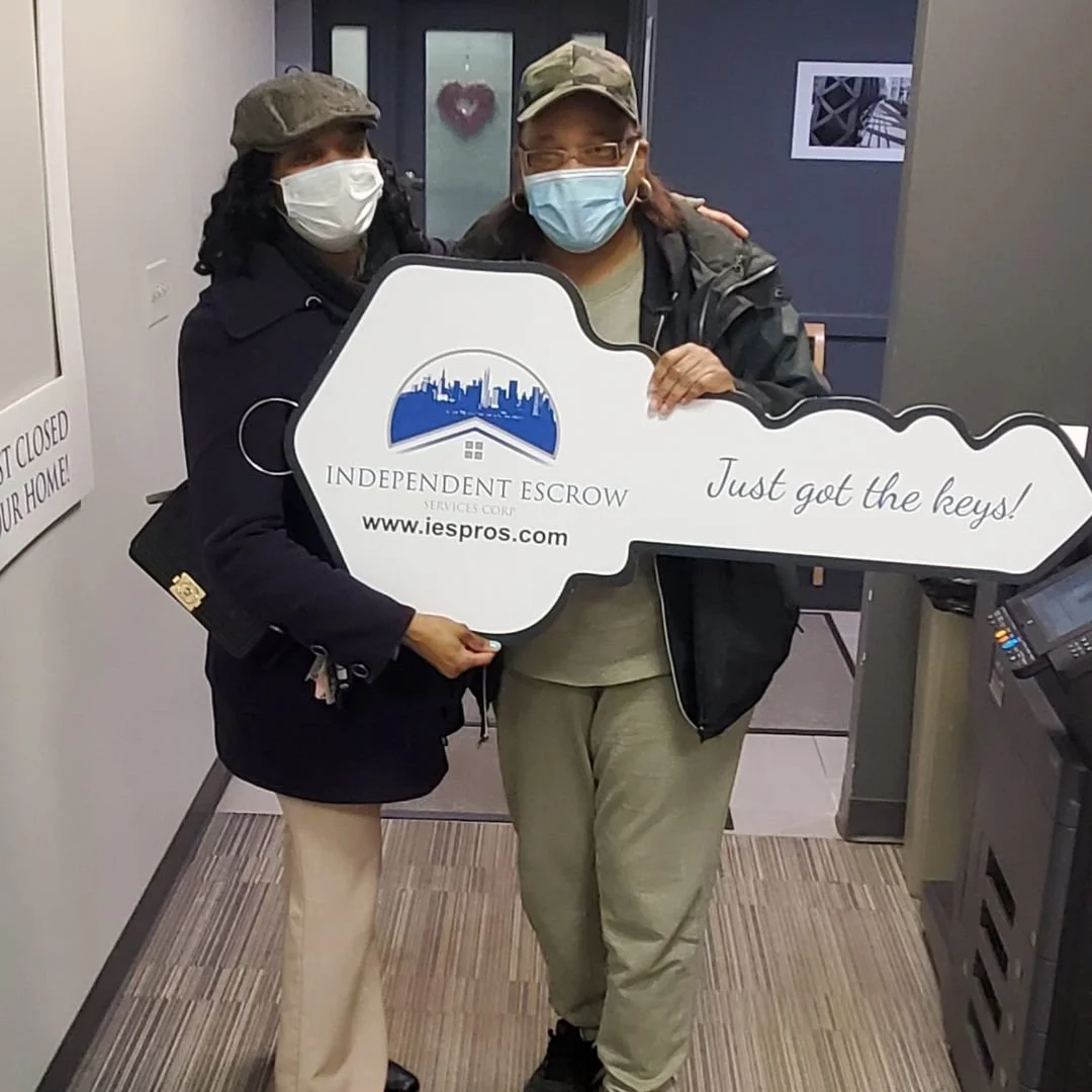 Two women wearing masks, holding a large key-shaped sign that reads 'INDEPENDENT ESCROW SERVICES CORP www.iespros.com Just got the keys!' inside an office building.