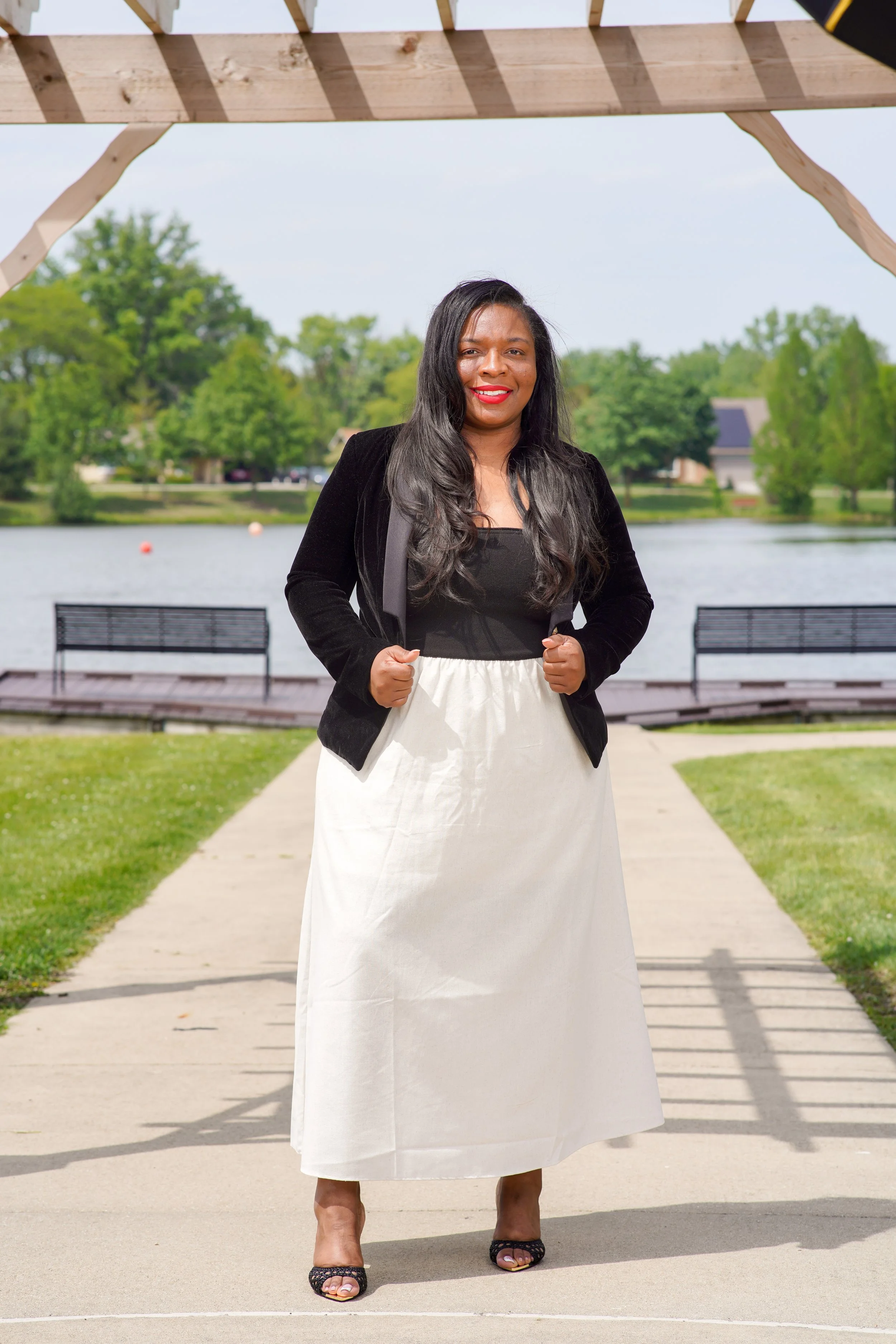 A woman with long, dark hair and red lipstick, wearing a black blazer, white skirt, and black open-toe shoes, standing outdoors near a lake with green trees and houses in the background, under a wooden structure.